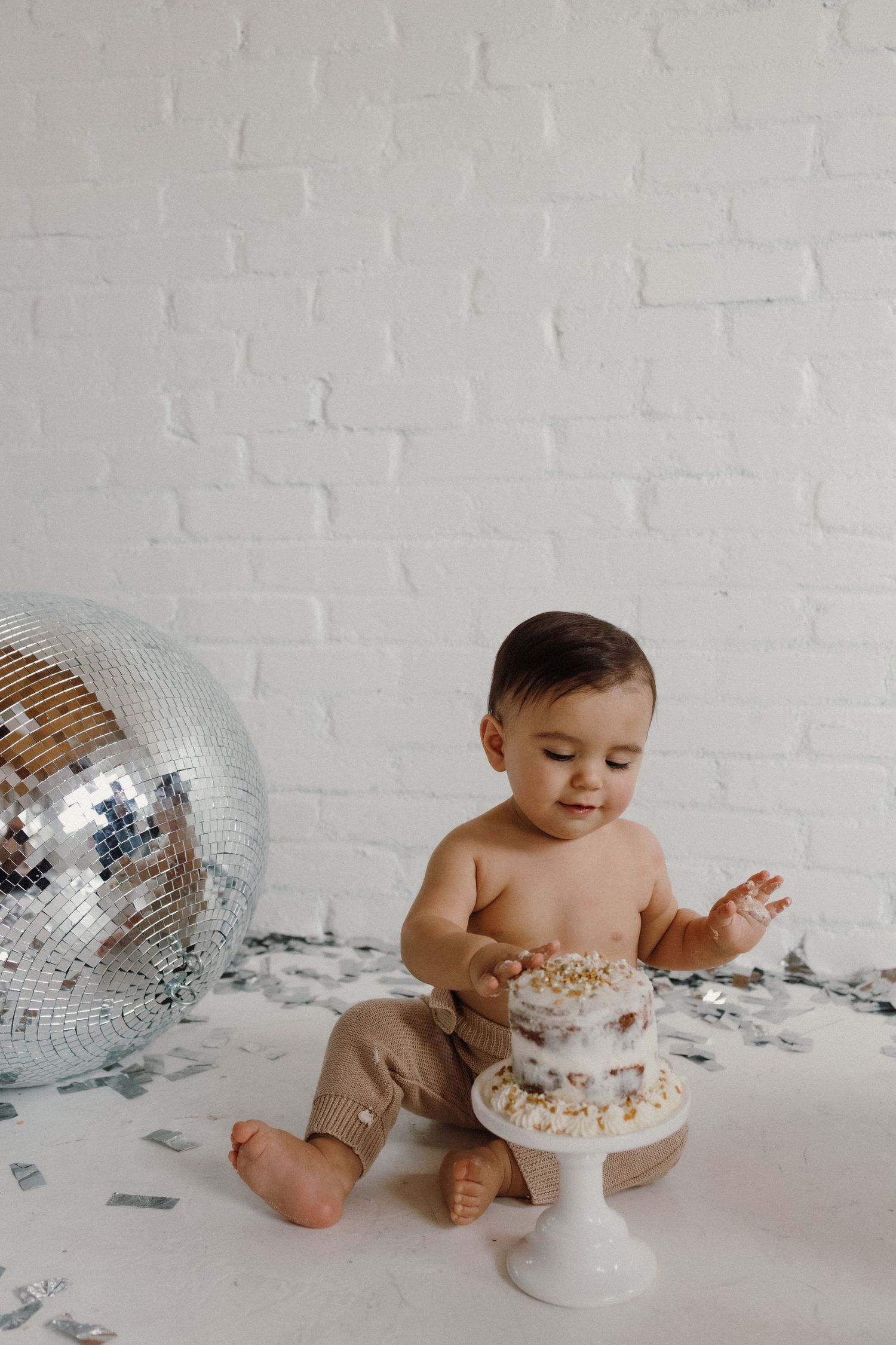 Baby in beige pants examining small white cake with disco ball decoration.