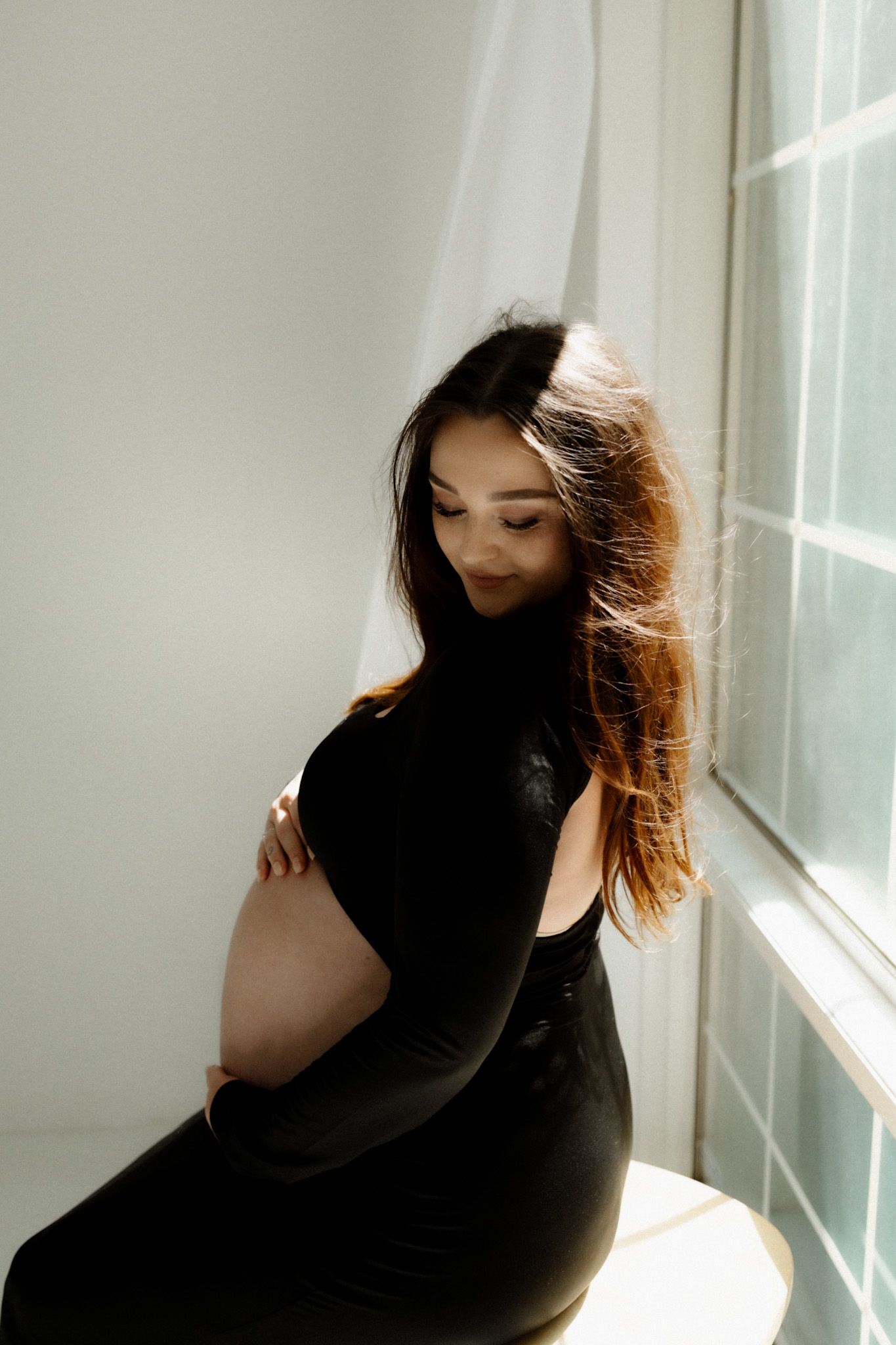 Elegant maternity portrait with sunlit window highlighting pregnant belly in black dress.