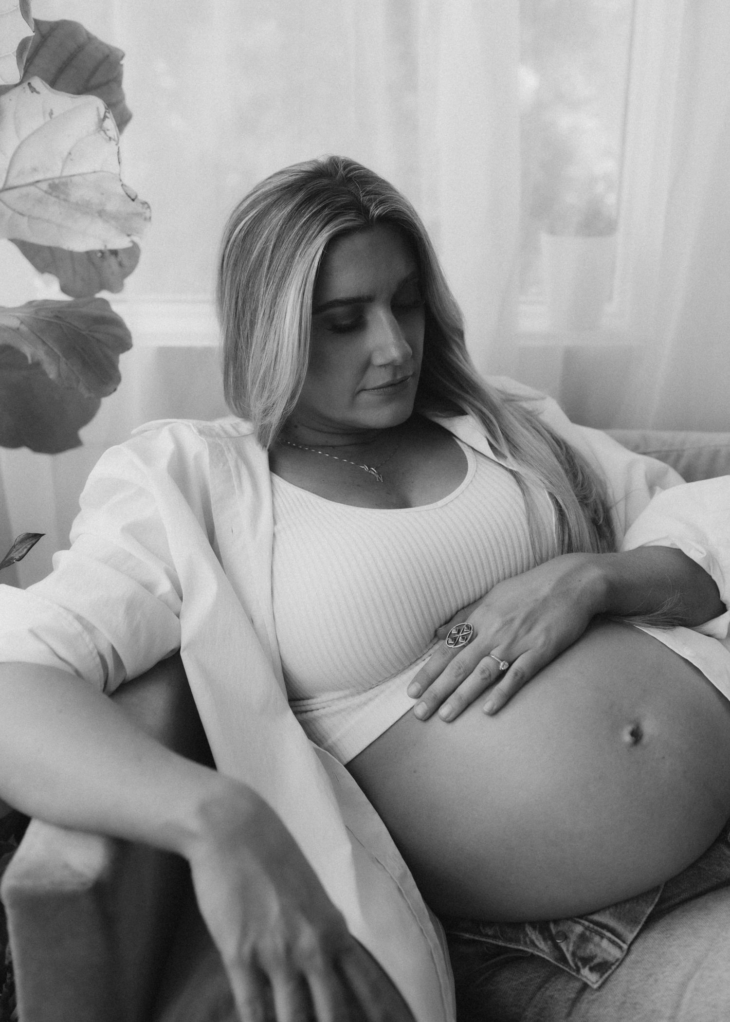 Black and white maternity photo showing an expectant mother in a white sweater resting on a couch in soft window light.