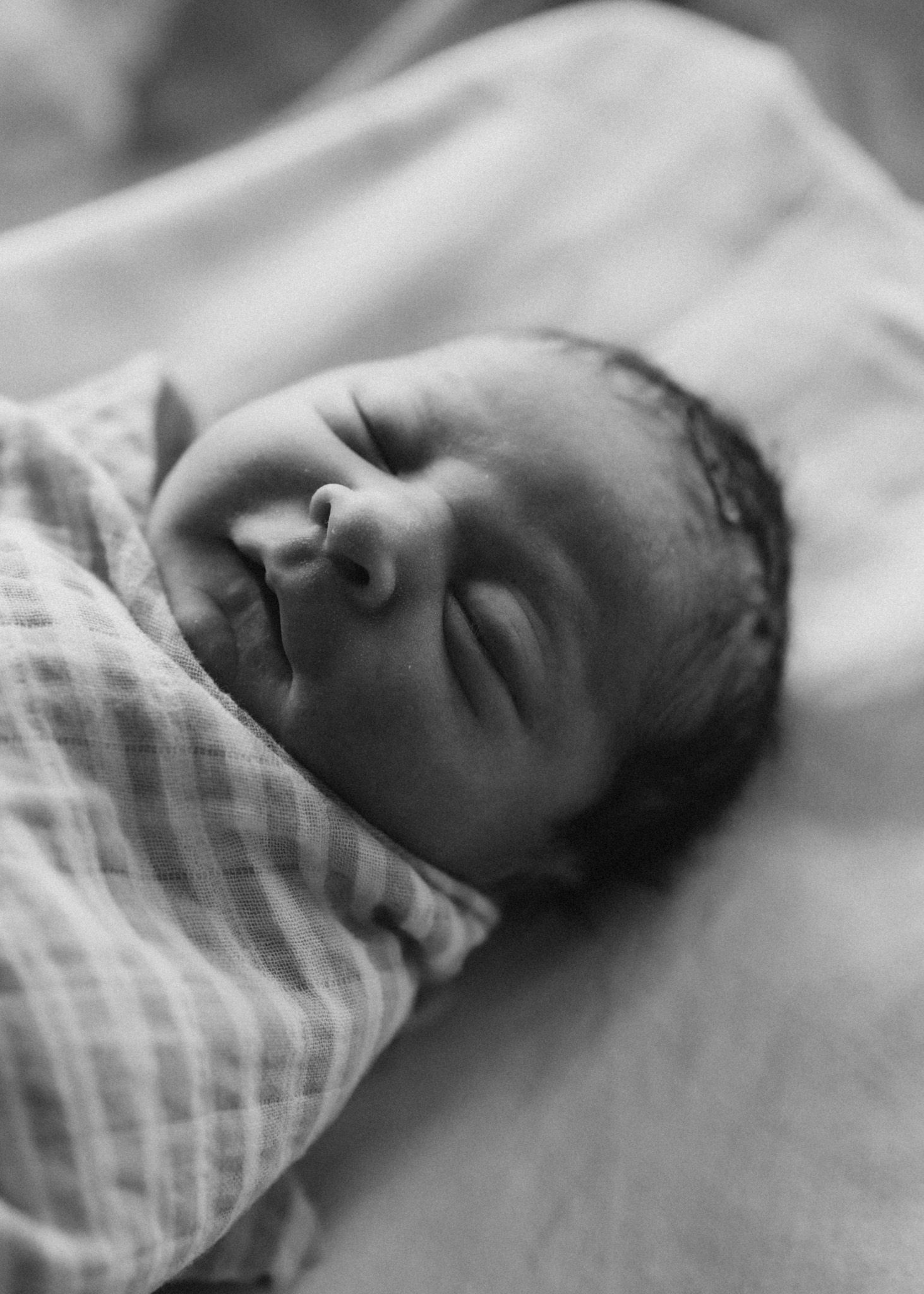 Close-up black and white photo of a sleeping newborn baby wrapped in a checkered blanket.