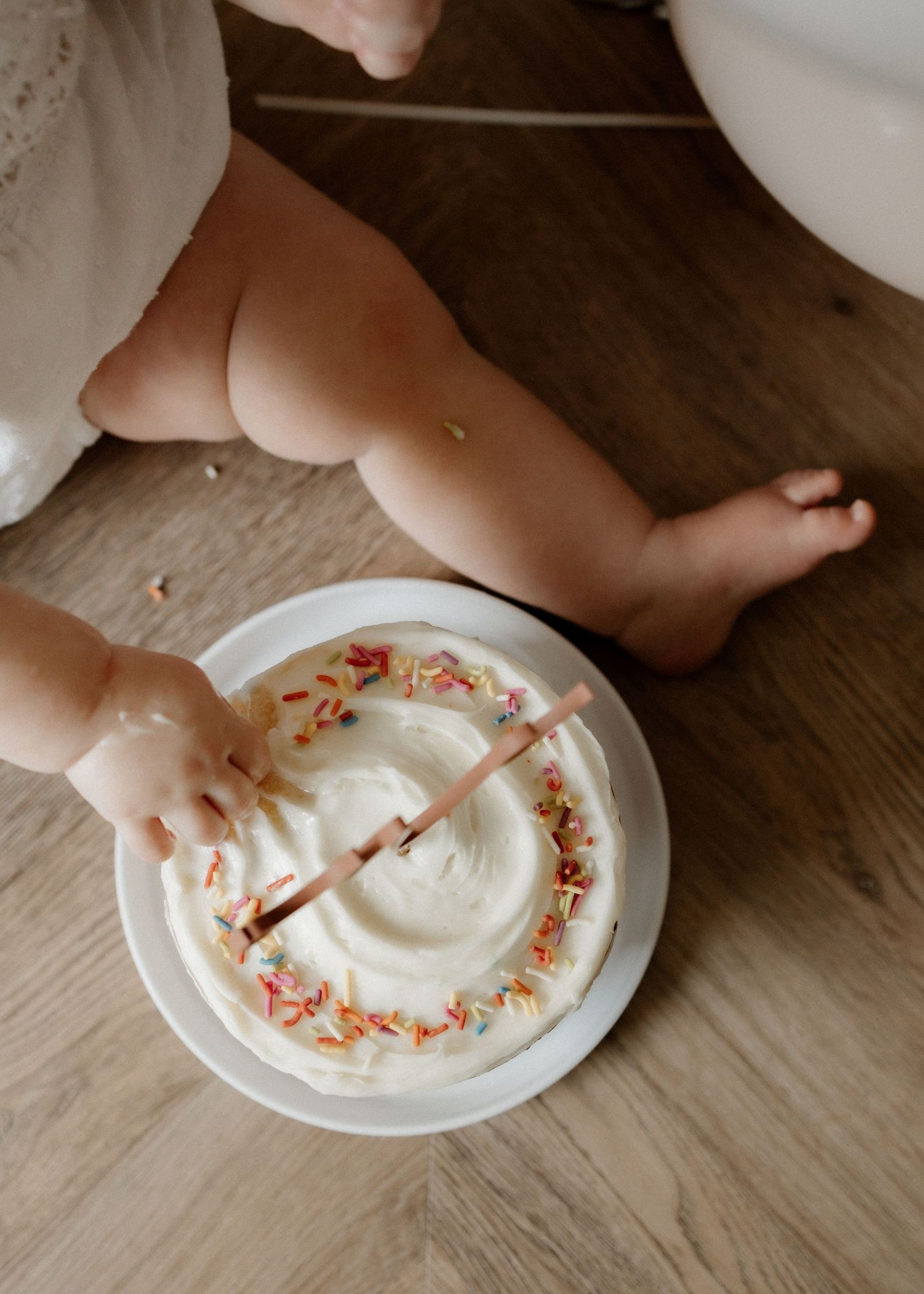 Close-up of tiny hands touching white frosted birthday cake on wooden floor.