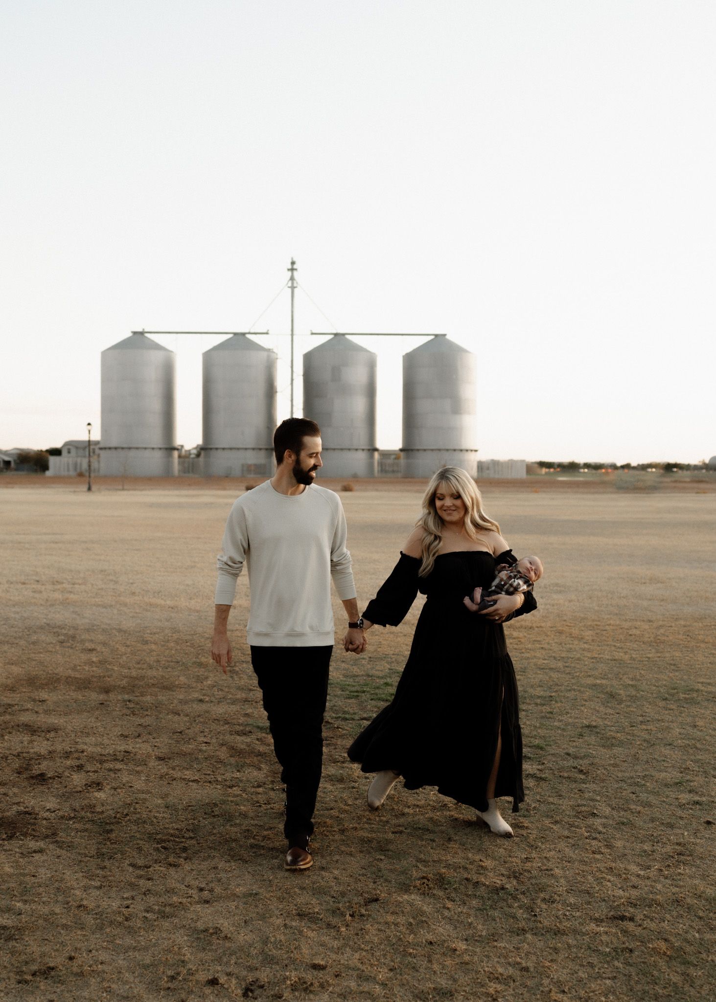 A couple walks together across an open field with grain silos in the distance.