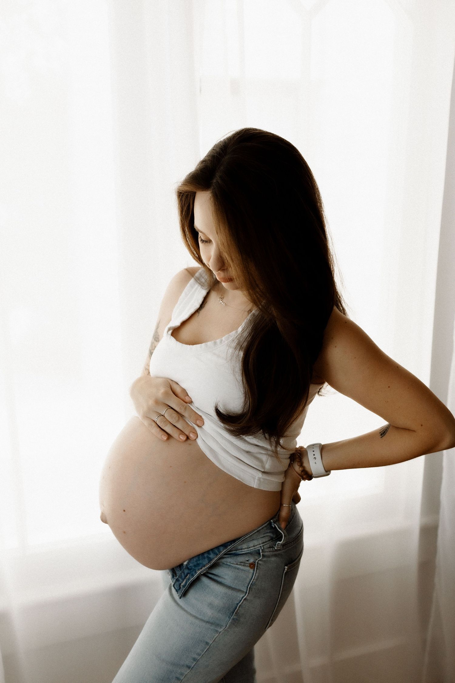 Side view of pregnant woman in white tank top and jeans by window.