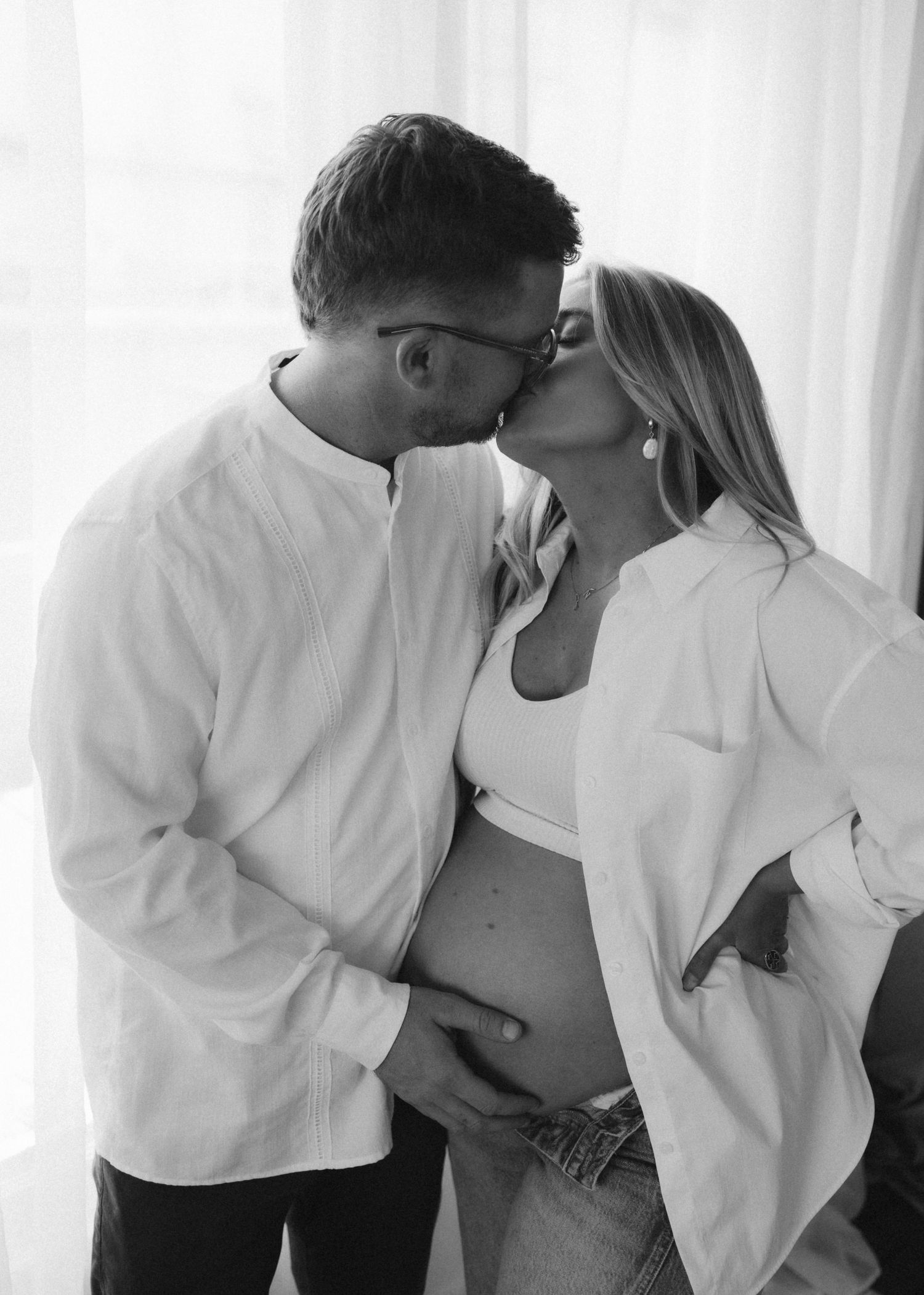 Black and white photo of a couple sharing an intimate moment during a pregnancy photoshoot, wearing matching white tops.