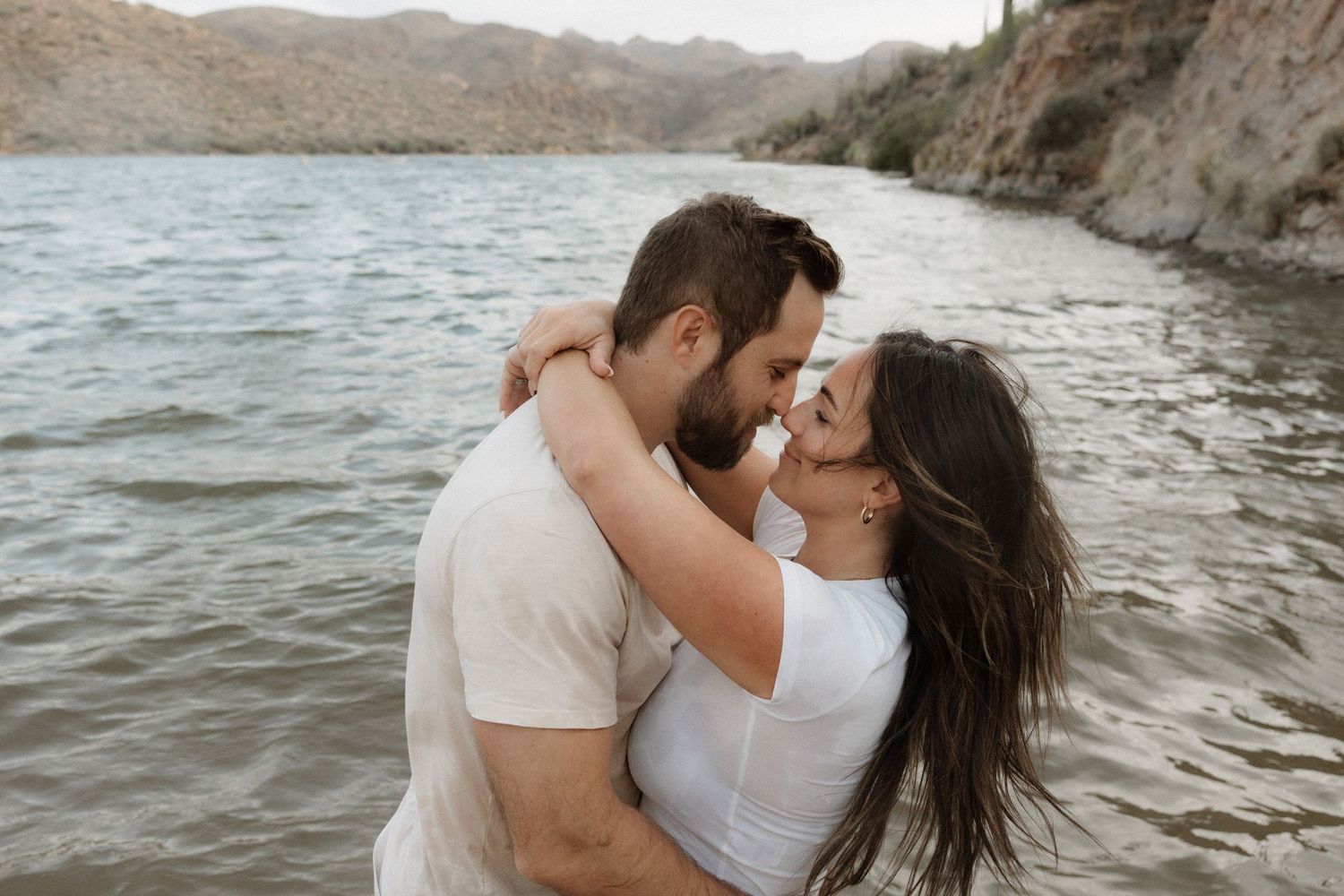 Two people embrace and kiss on a rocky beach with waves crashing around them in a romantic coastal photoshoot.