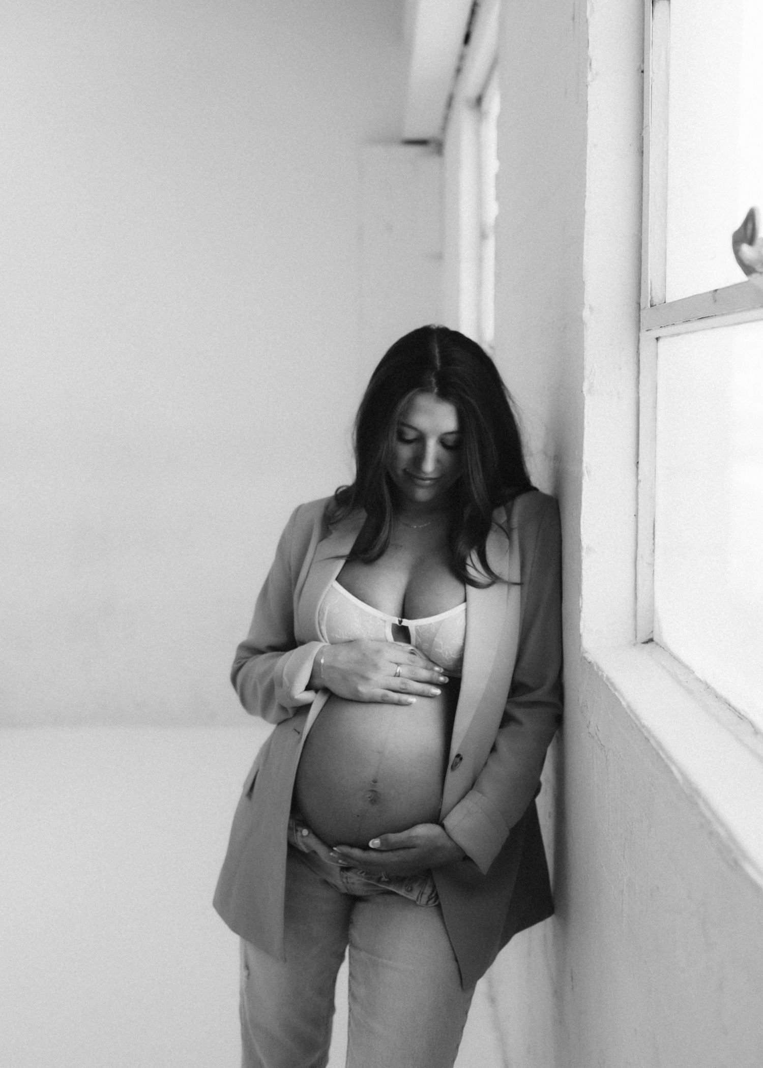 A thoughtful black and white portrait of a pregnant woman in business attire standing by a window.