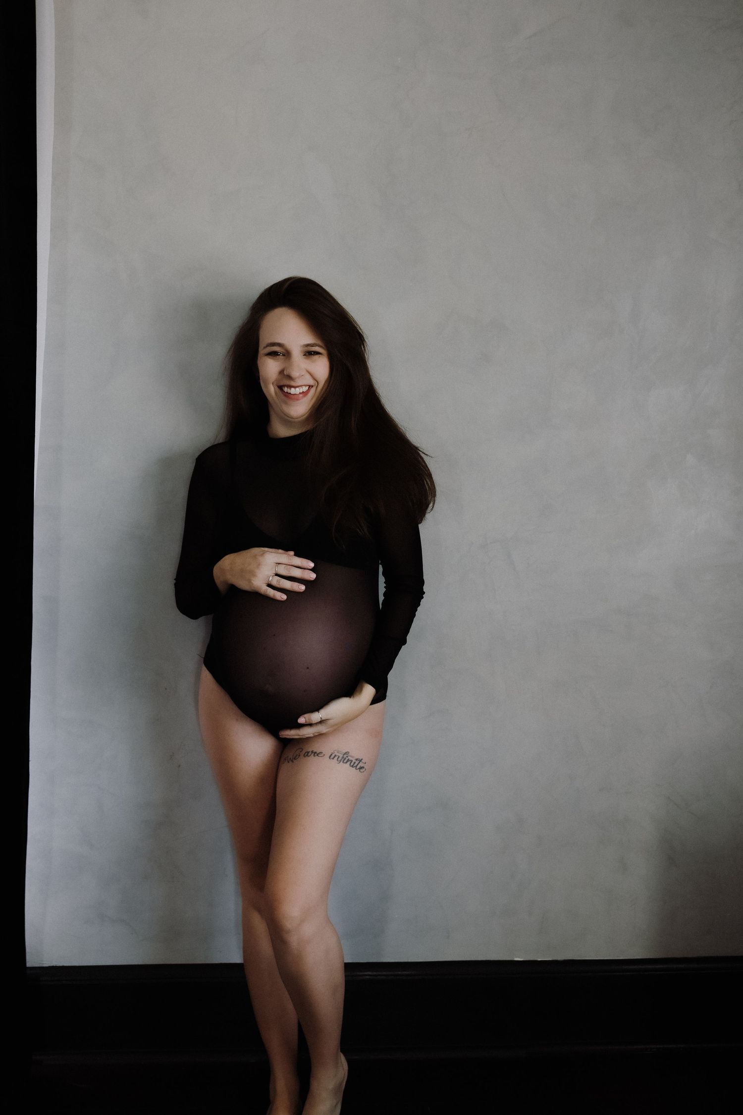 Intimate maternity portrait showing a person in black against a minimalist gray wall.