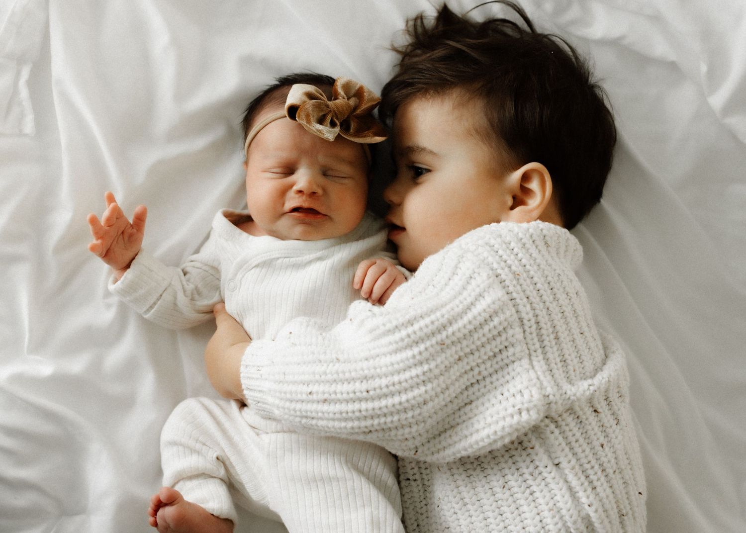 Two children in white sweaters lying together on white bedding while sharing a tender moment.