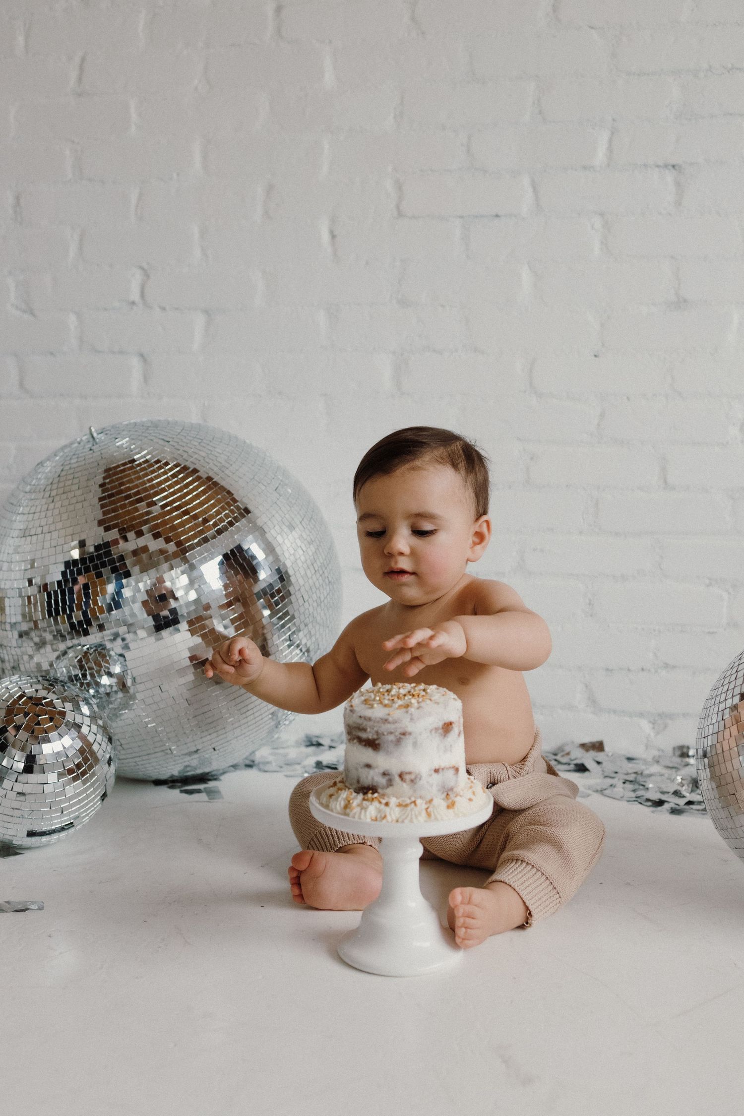 Baby sitting next to disco ball and small white cake on stand against brick wall.