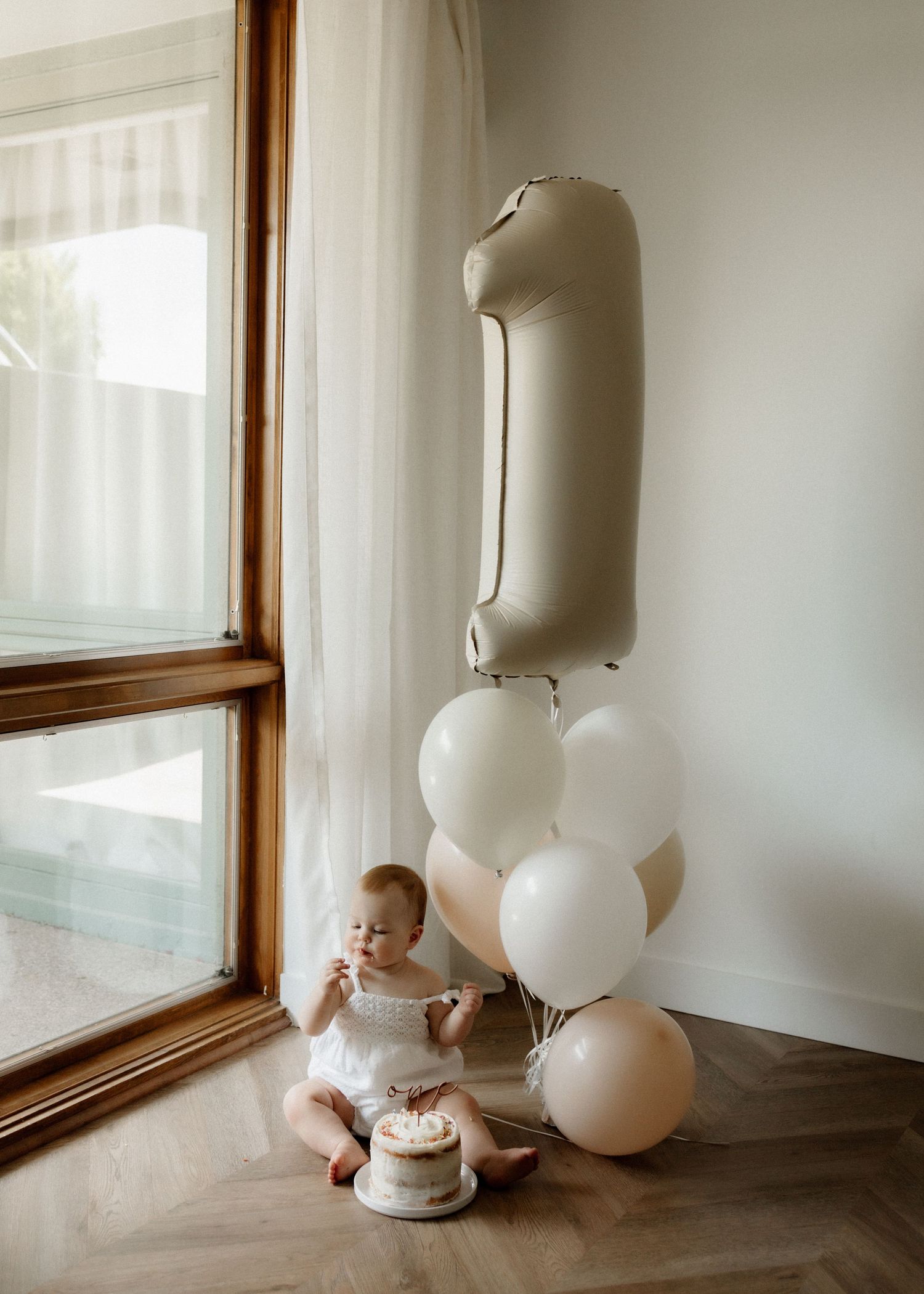 Baby in white dress sits by window with first birthday cake and neutral balloons.