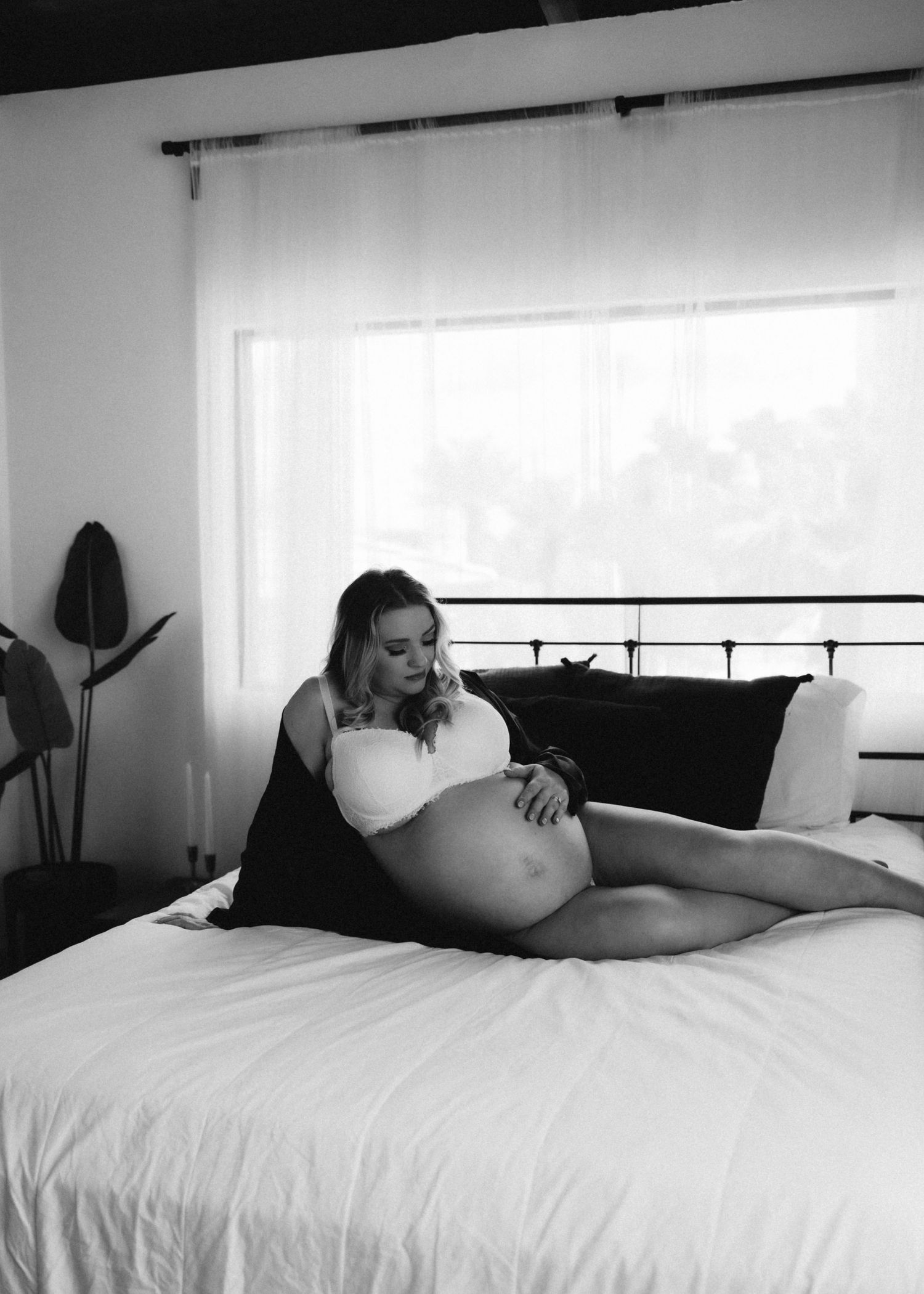 Black and white maternity boudoir photo on bed with natural window lighting.