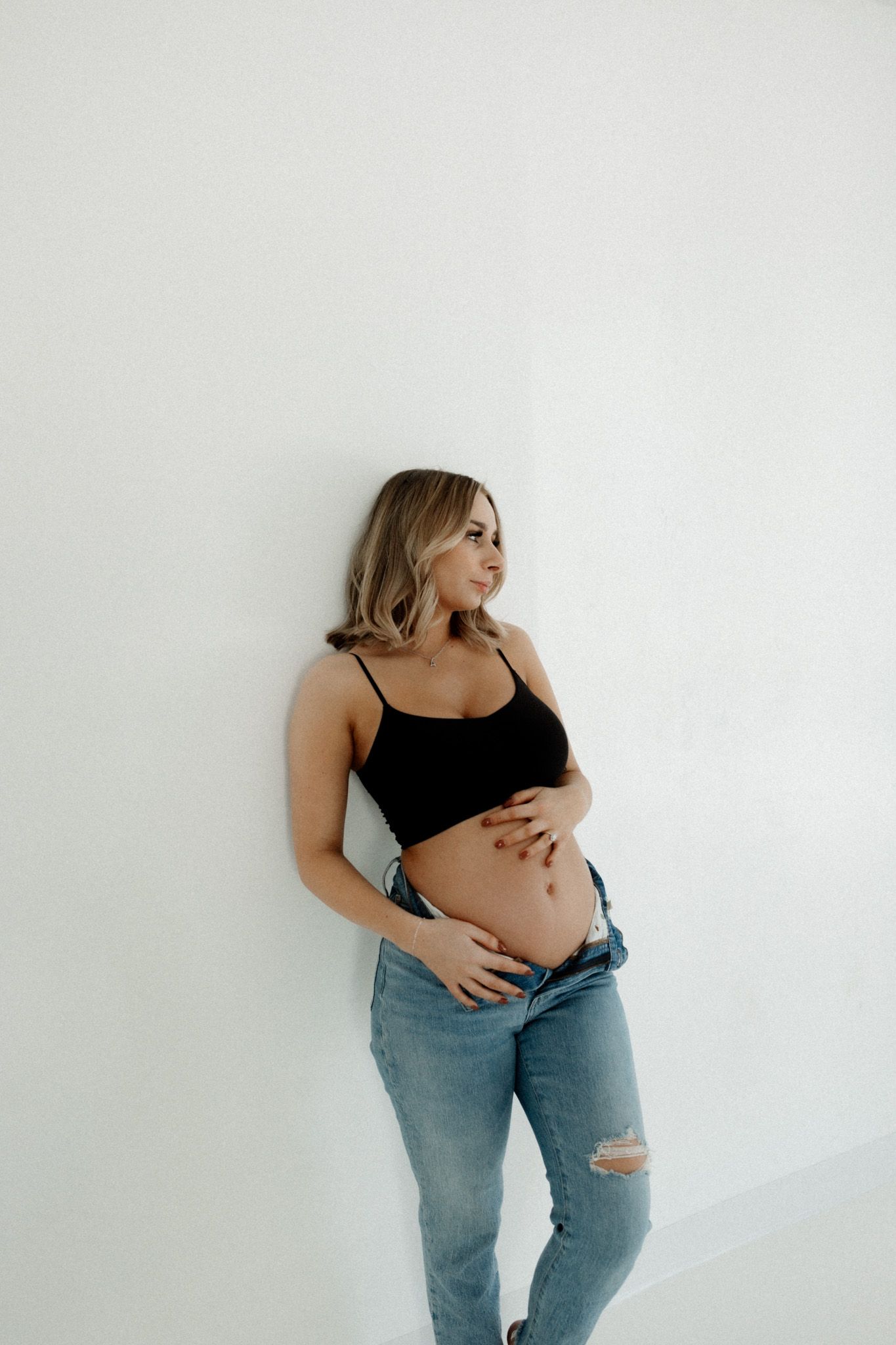 Casual maternity photo in black top and distressed jeans against white wall.