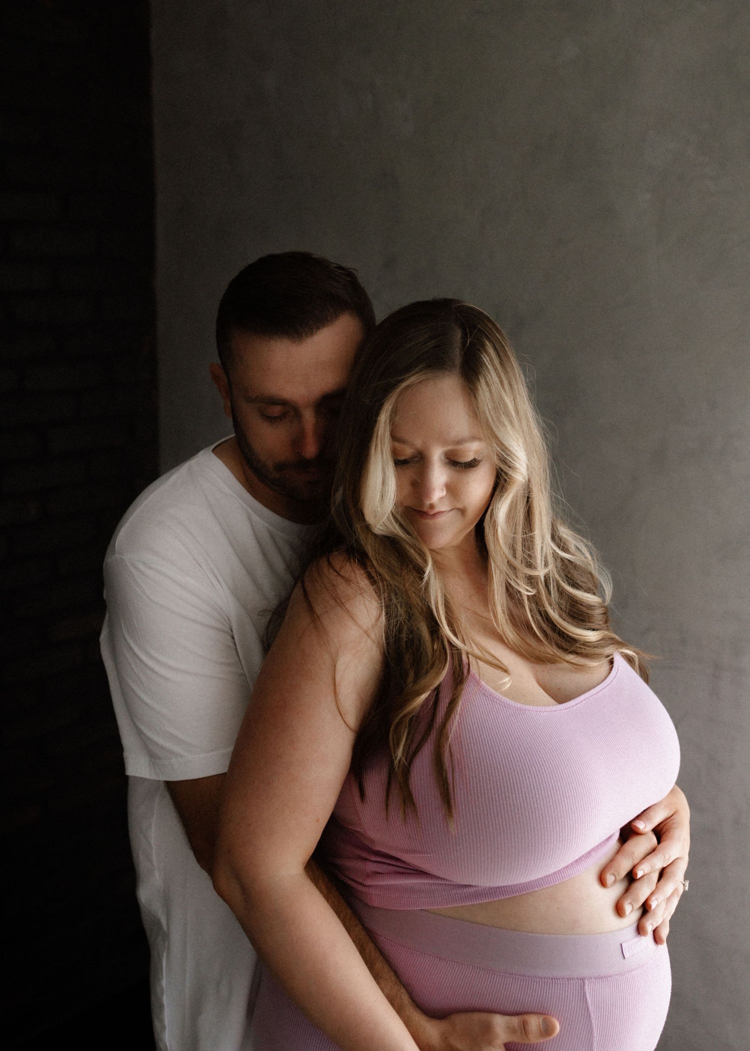 A tender maternity photo of a couple embracing in soft lighting against a grey wall.