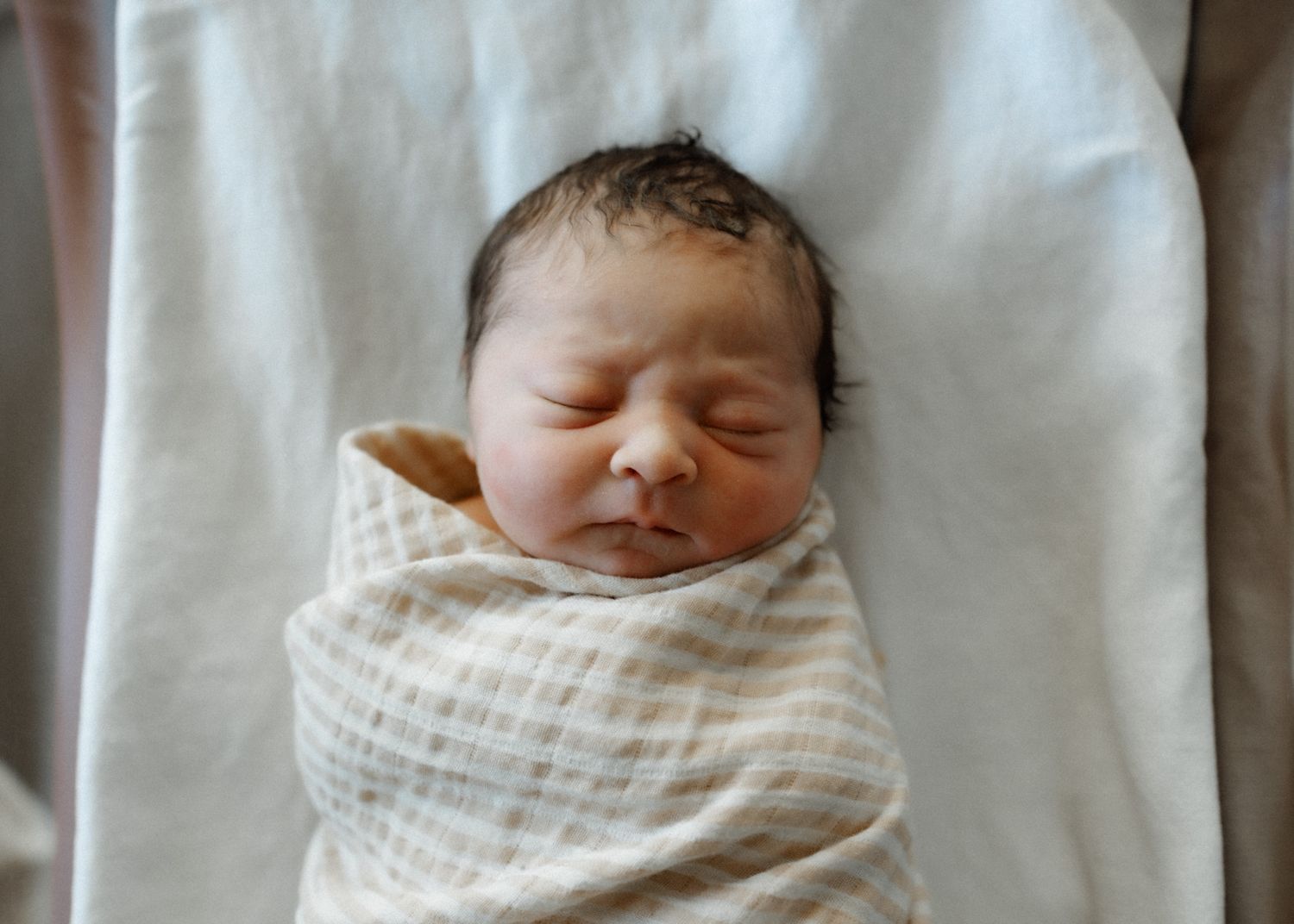 Peaceful newborn baby swaddled in a white checkered blanket sleeping on white bedding.