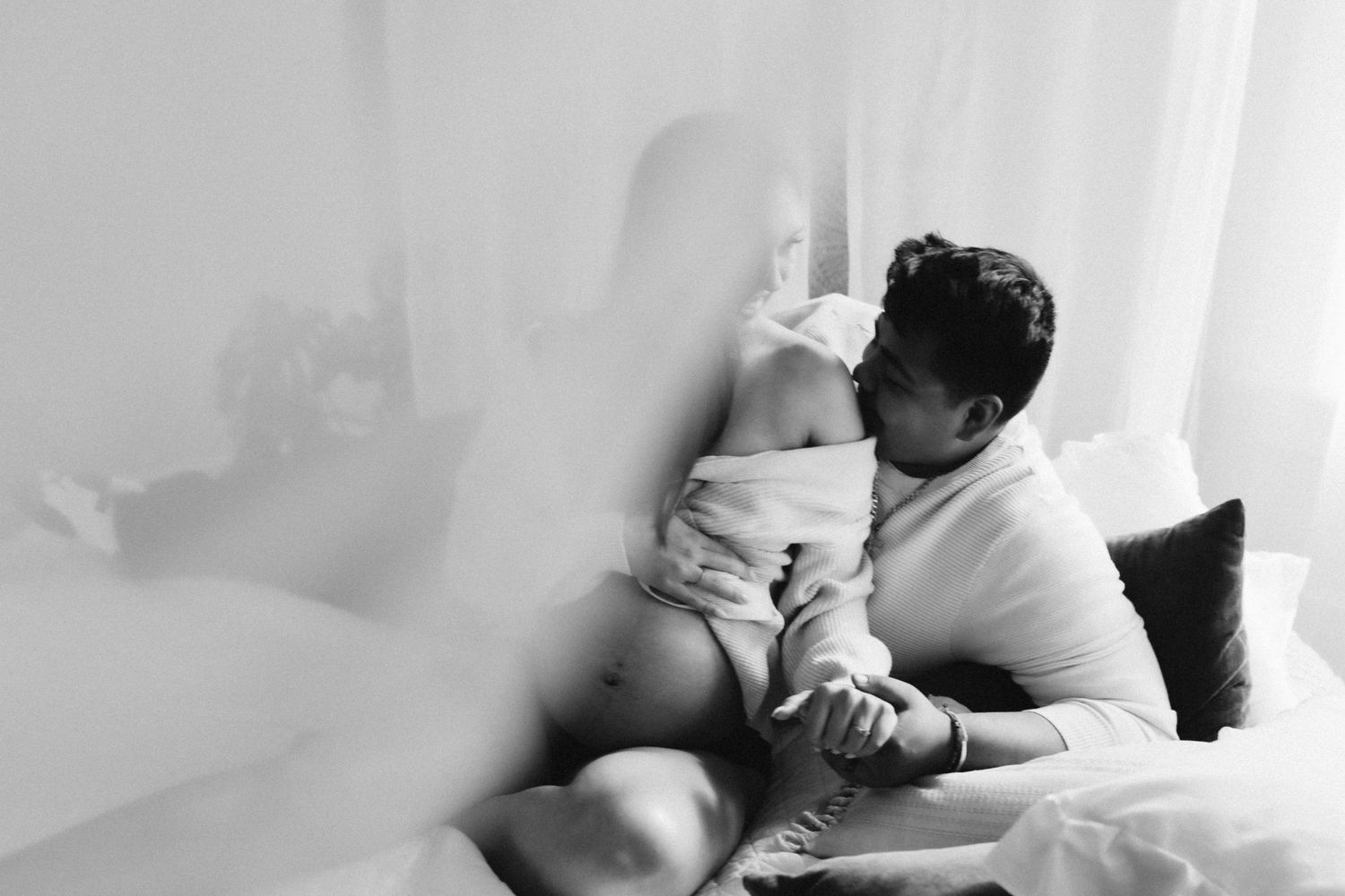 Gentle black and white indoor maternity moment captured through sheer curtains.