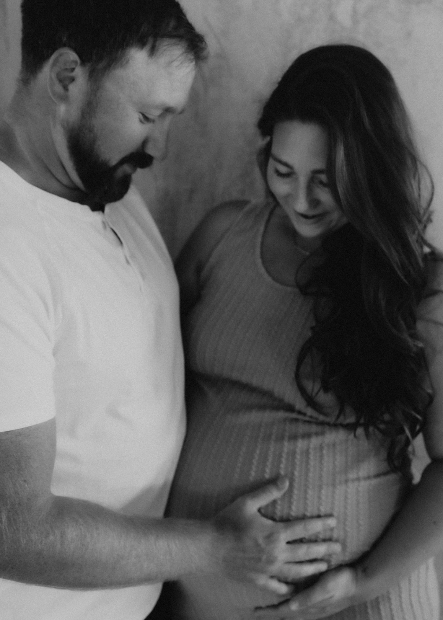 A black and white pregnancy photo showing a couple looking lovingly at her belly in an intimate moment.