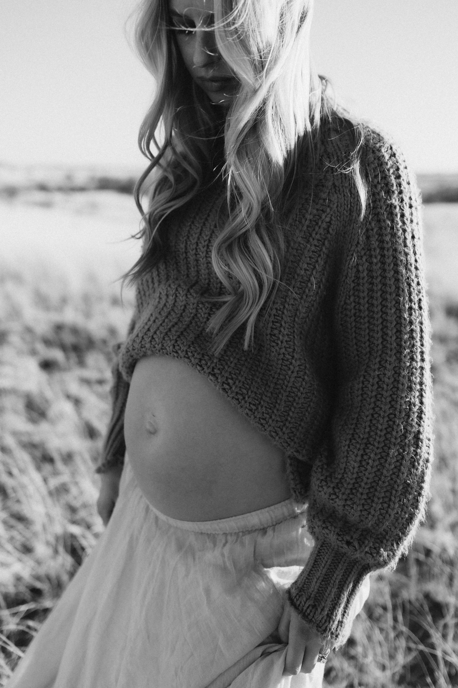 Black and white photo of a person wearing a cropped knit sweater and flowy white dress in a pastoral outdoor setting.