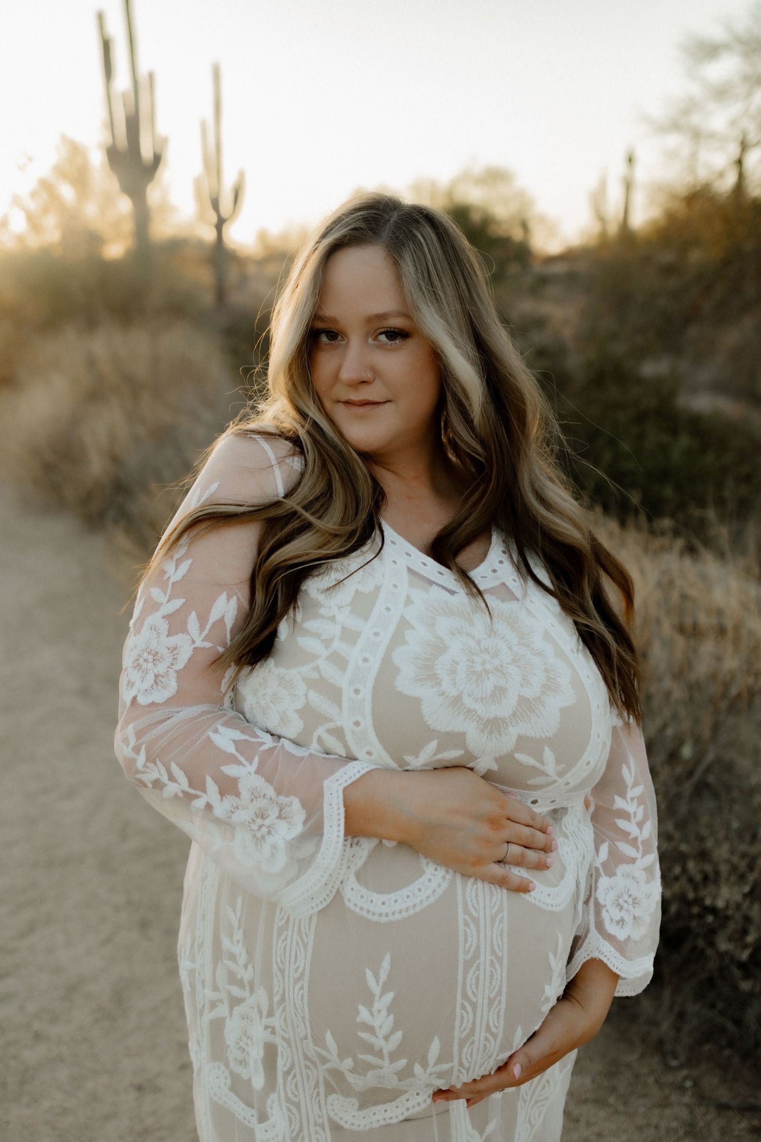 Woman in lace maternity dress poses against desert landscape at sunset.