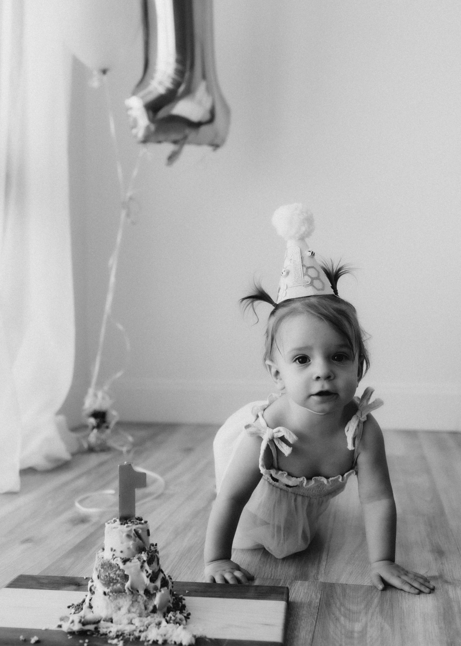 Toddler in white dress crawls near birthday cake with silver balloon in minimalist black and white photo.