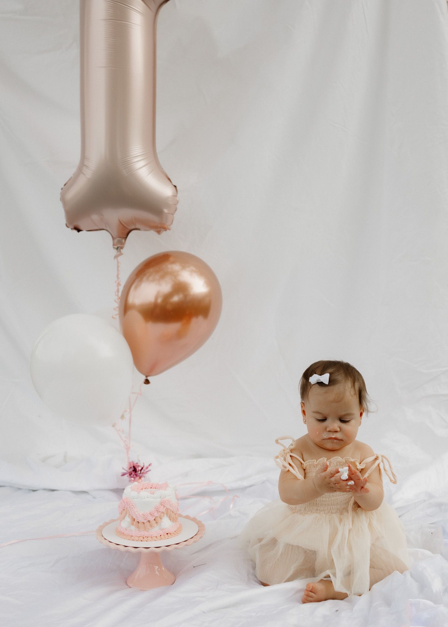 A baby in a tulle dress sits near rose gold and white balloons with a small cake for a birthday shoot.