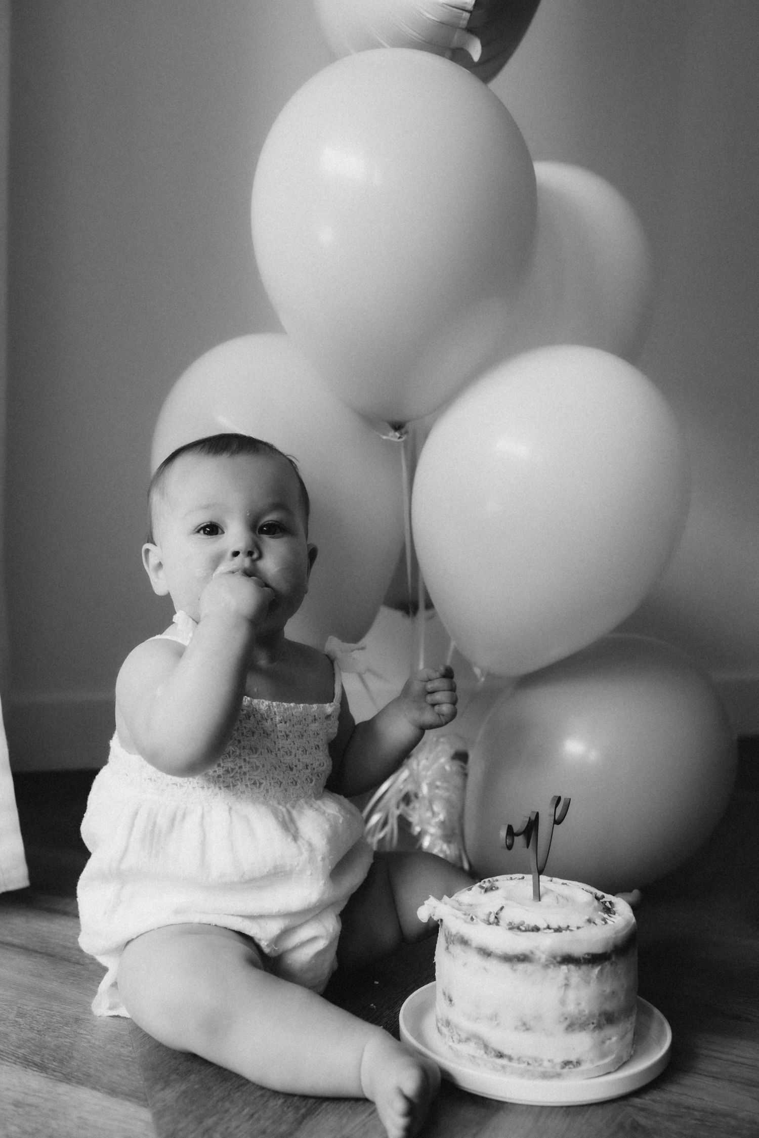 Baby in white outfit sits with birthday cake and balloons during milestone photo session.