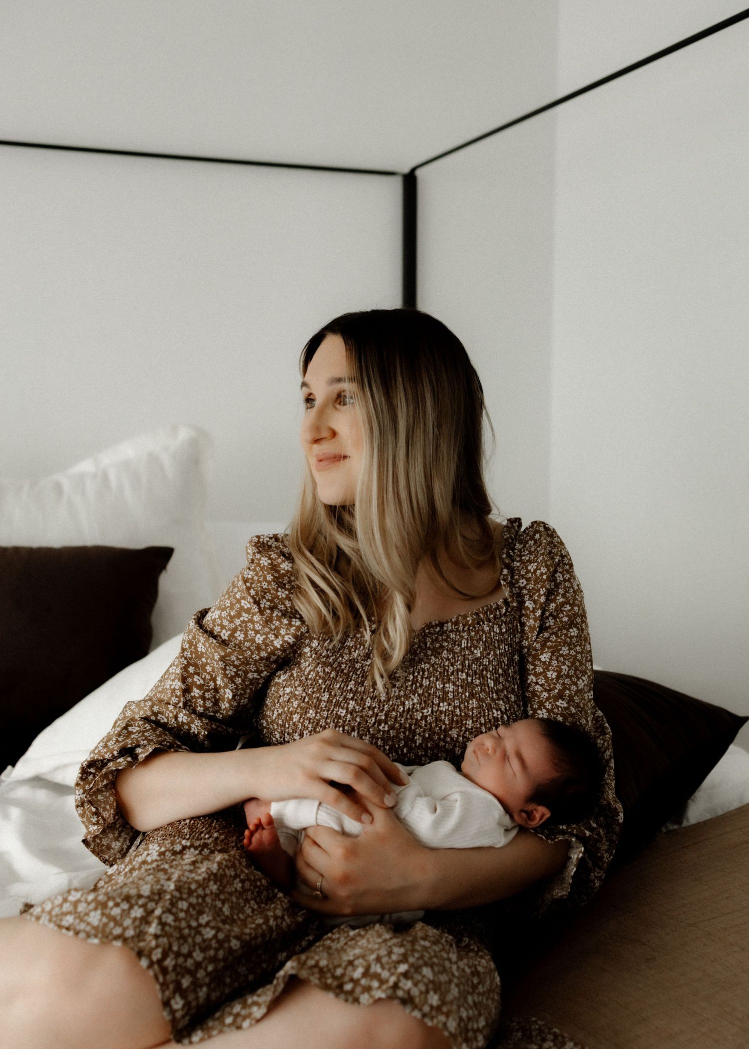 Parent in brown floral dress bottle feeding infant on couch.
