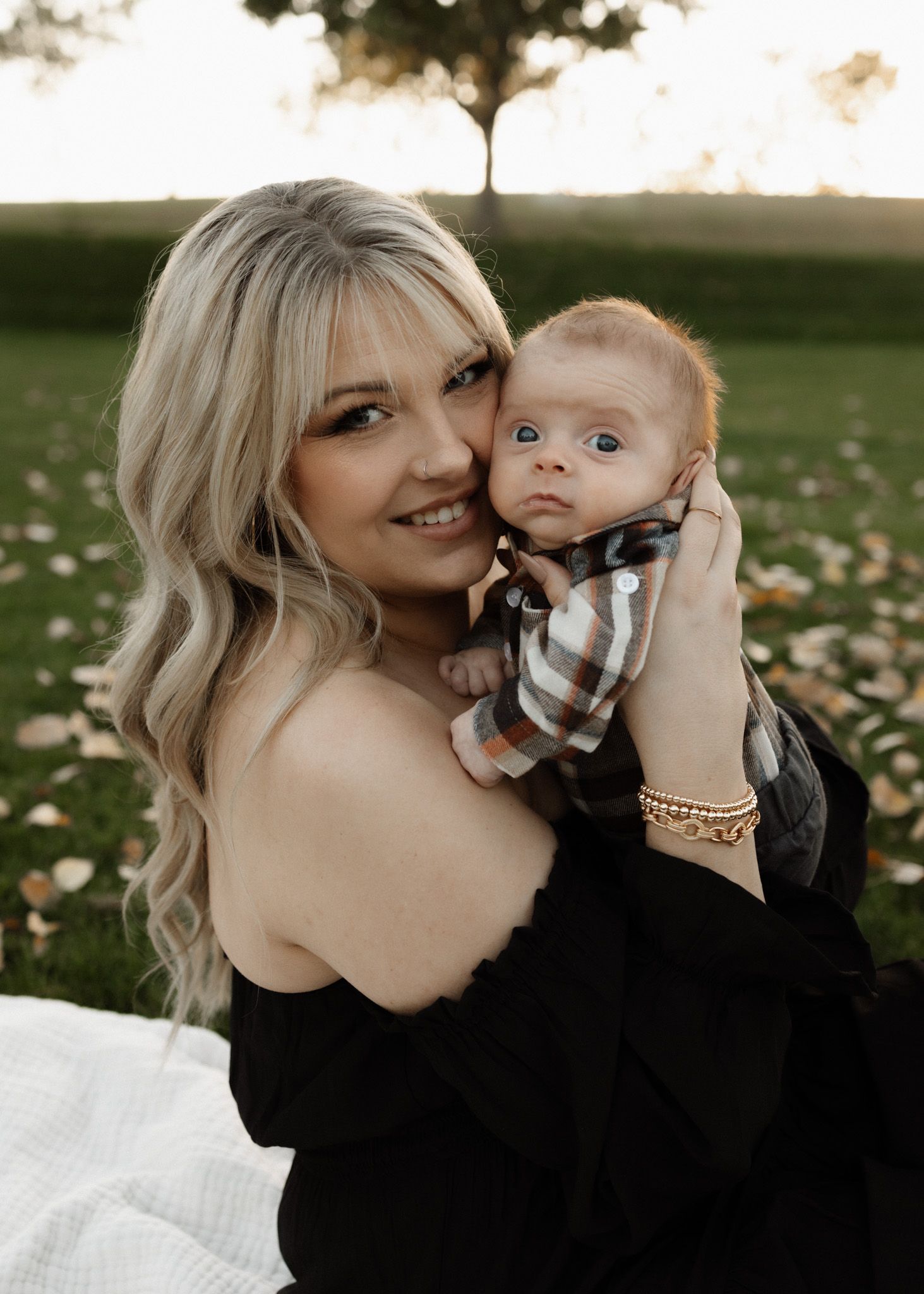 Mother in black dress holding baby in plaid shirt outdoors during sunset.