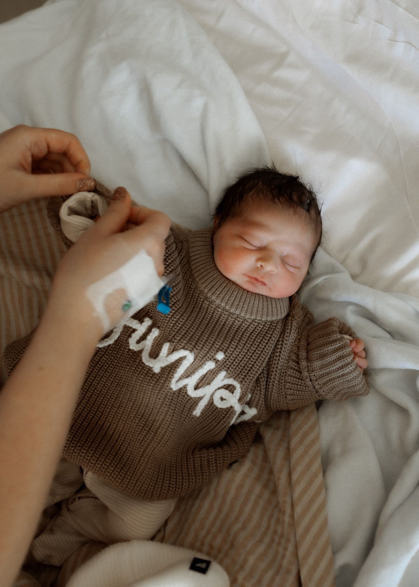 Peaceful sleeping newborn wearing brown knitted sweater rests on white bedding.