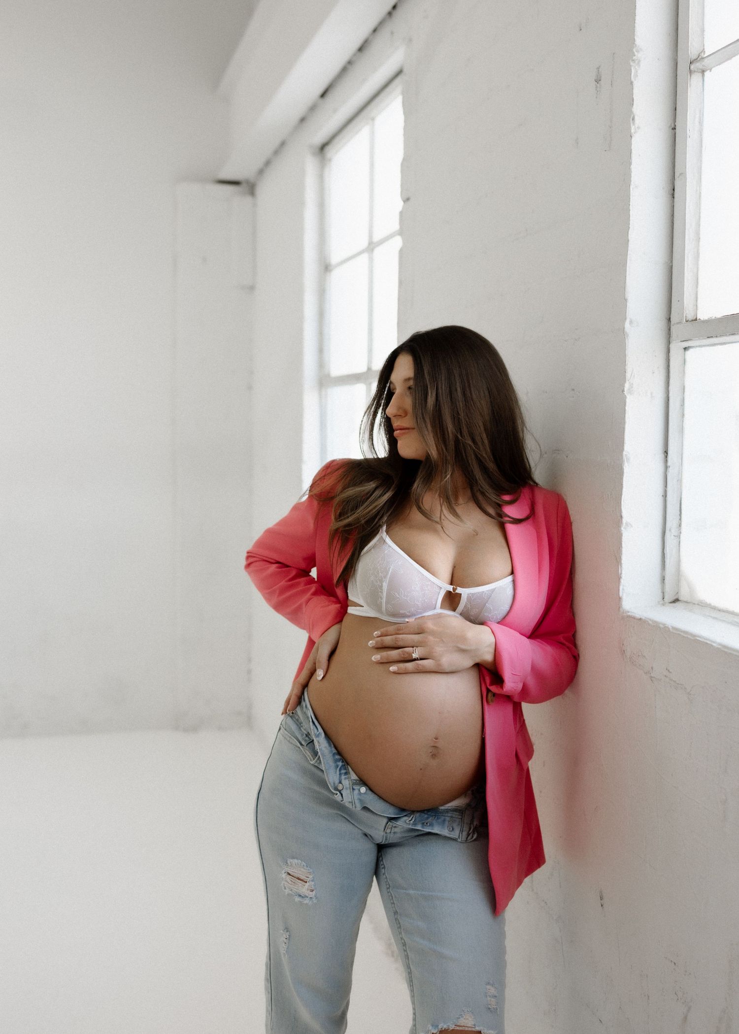 An expectant mother wearing a pink cardigan and distressed jeans stands by a bright window in a minimalist white room.