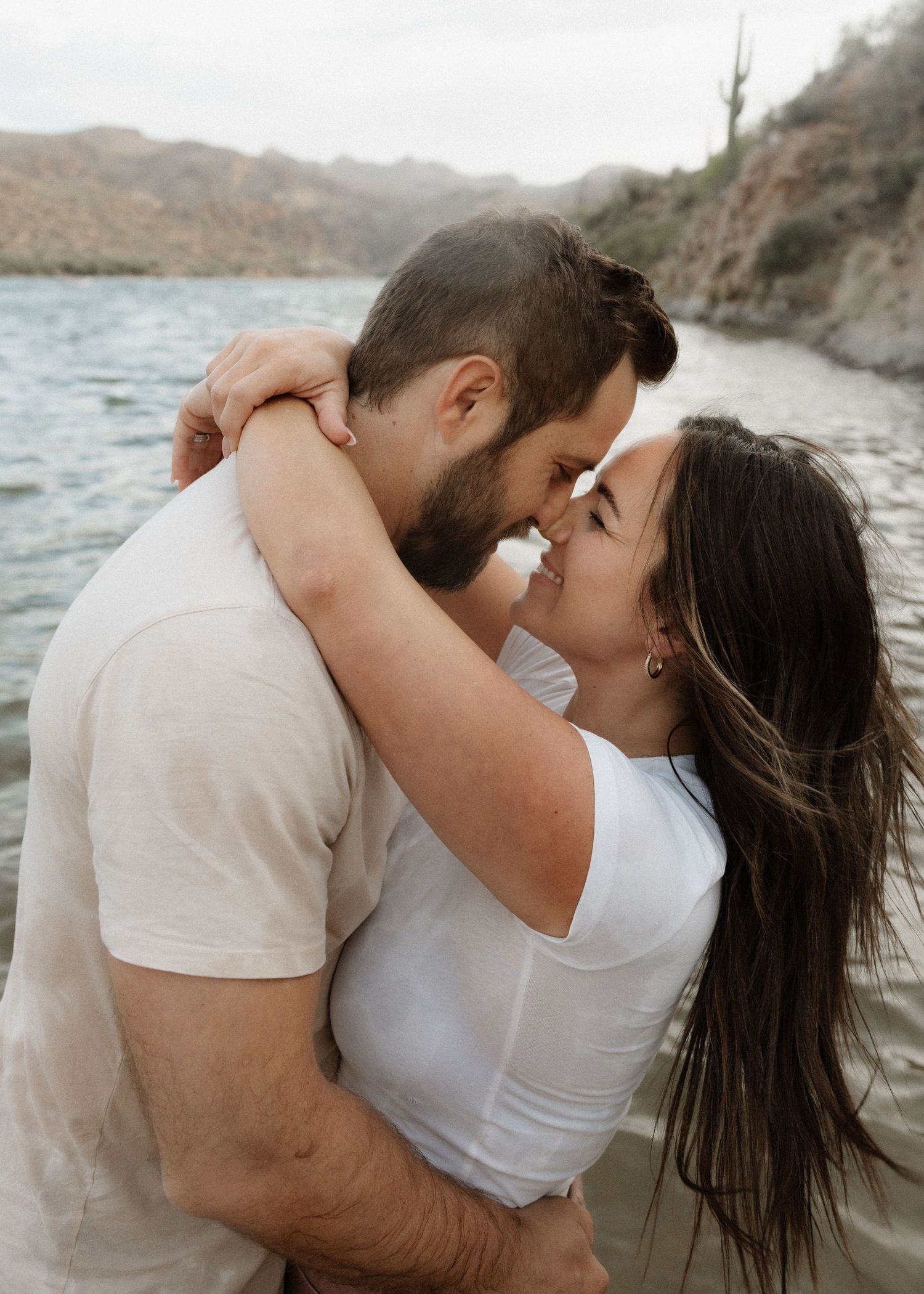 A romantic couple embraces near a rocky lake shore at sunset.