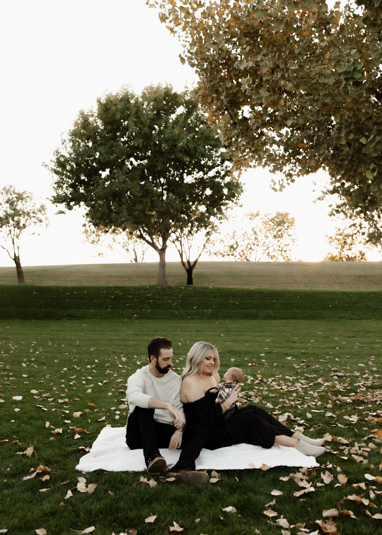 Family sitting on blanket surrounded by fallen leaves in park setting.