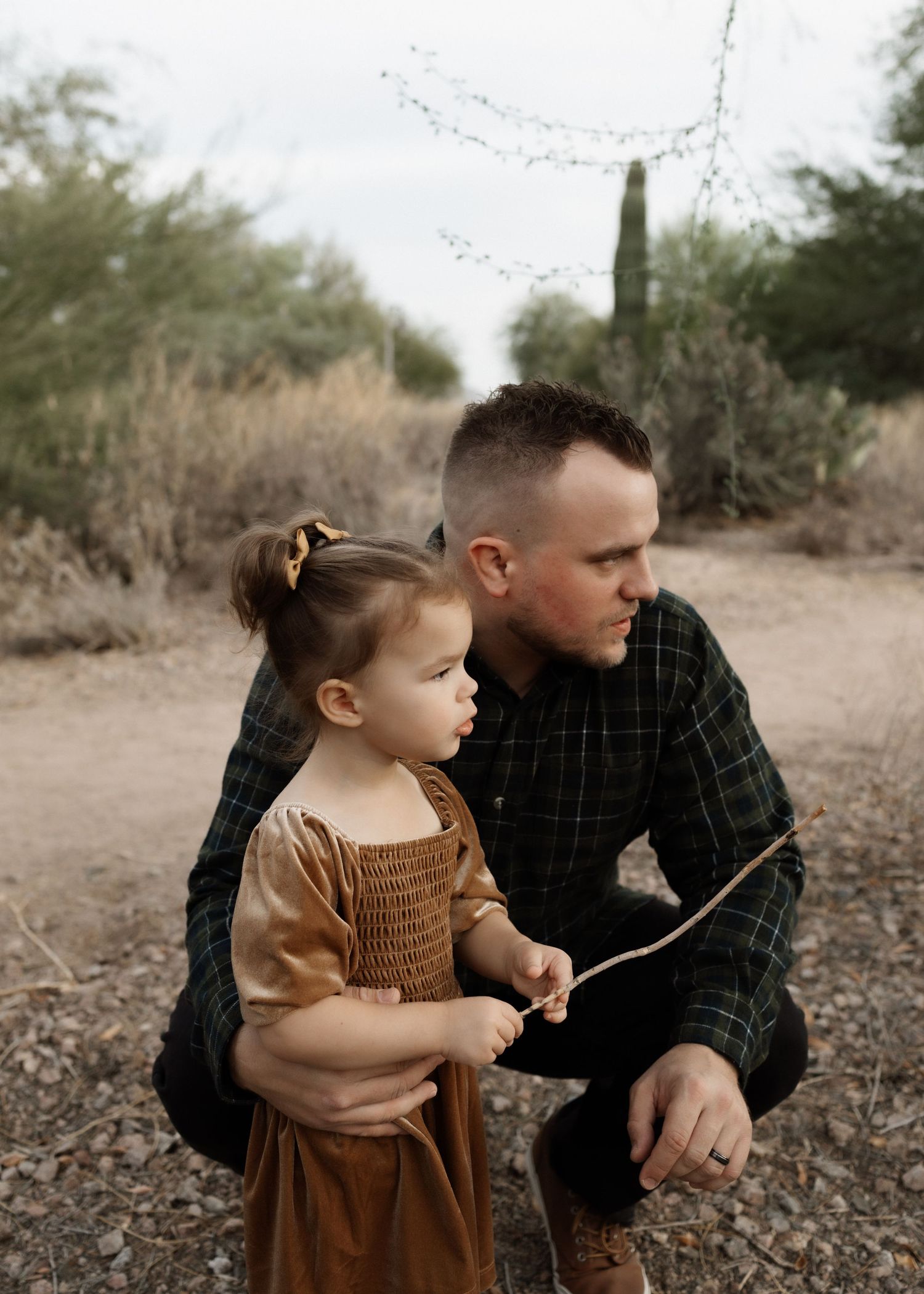 Adult in plaid shirt kneels beside small child holding a stick in a desert setting.