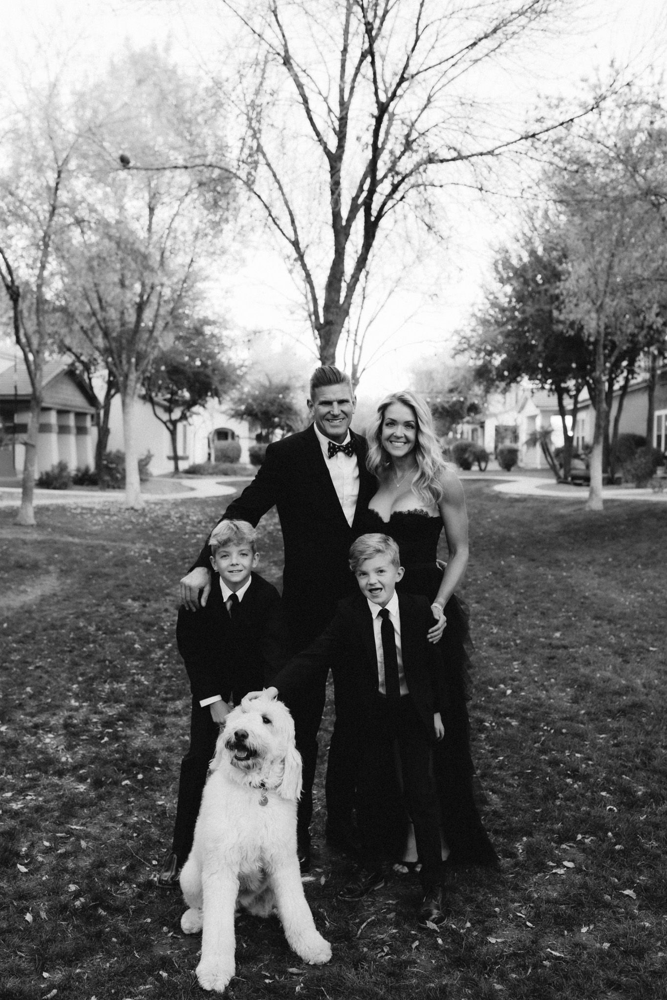 Family poses outdoors with their white fluffy dog wearing formal attire.
