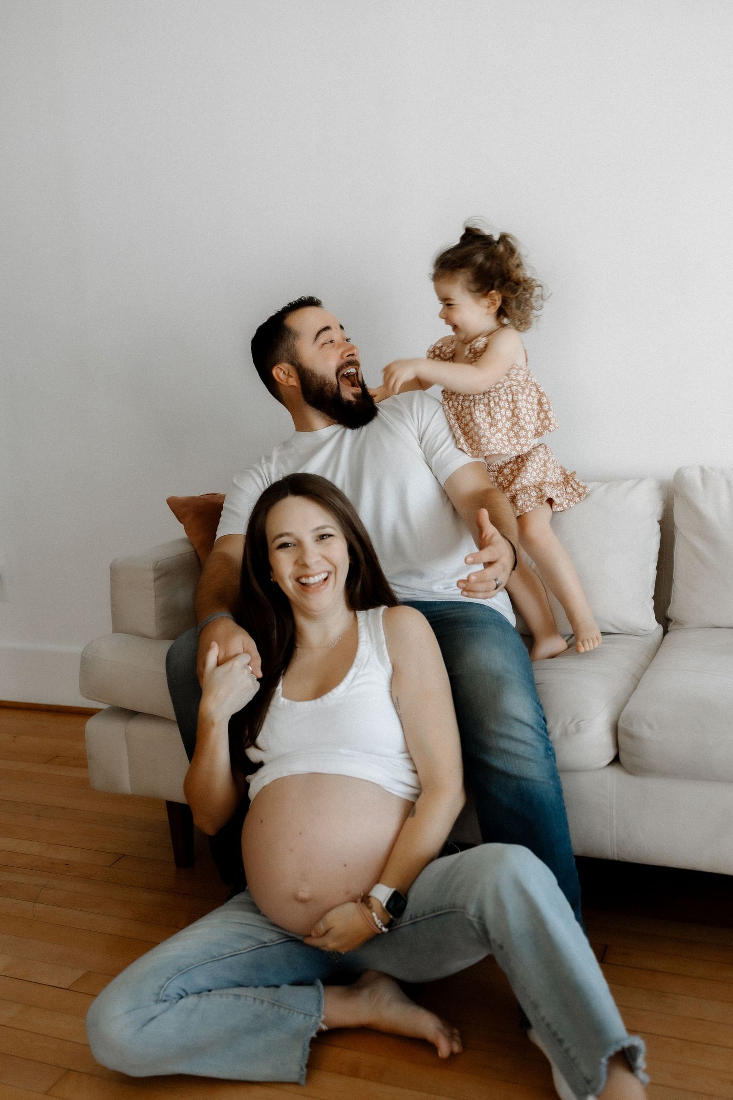 Family sitting together on floor by white couch with expectant mother in white top and jeans.