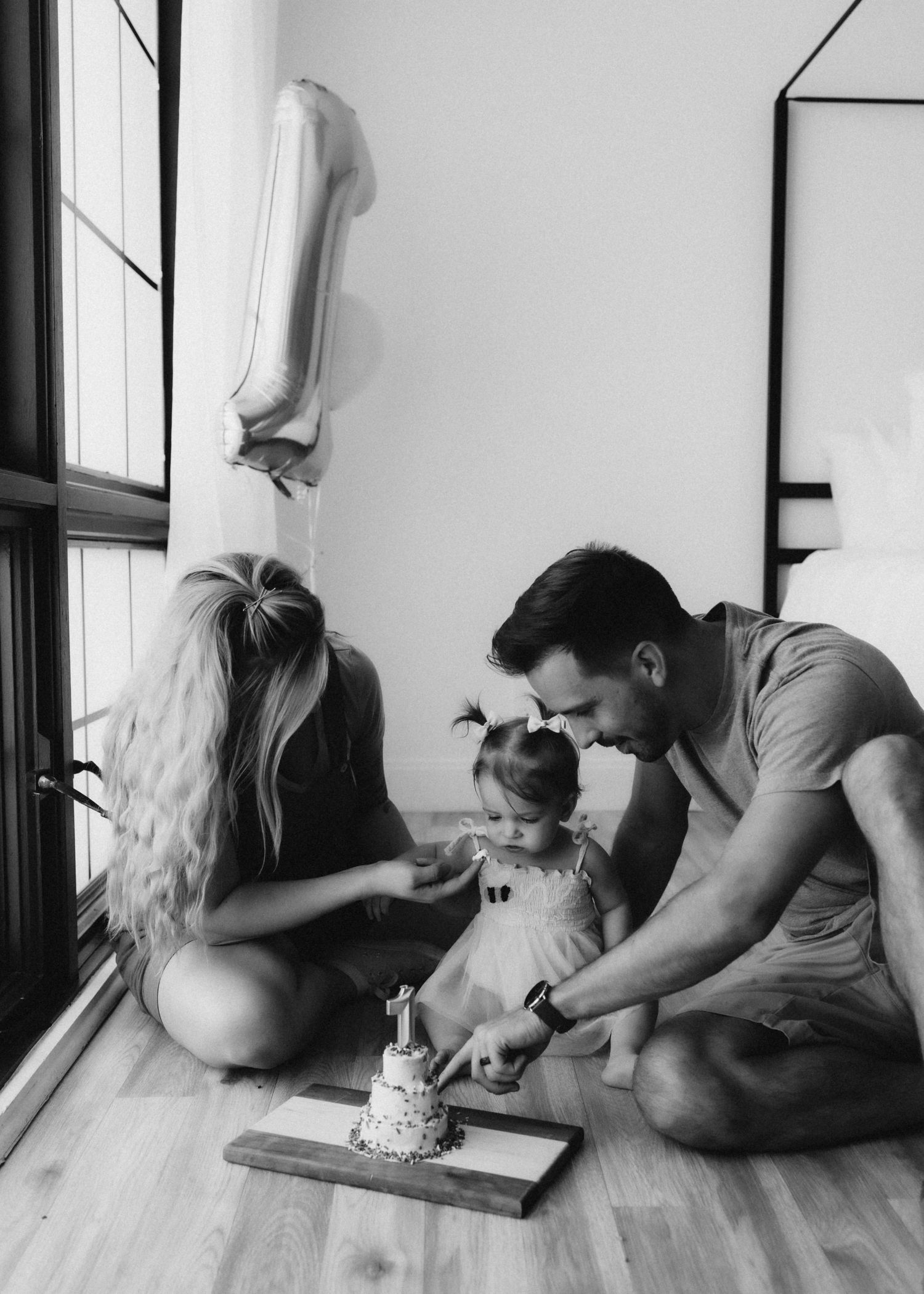 Family celebrates with small cake and silver number one balloon by window in black and white photo.