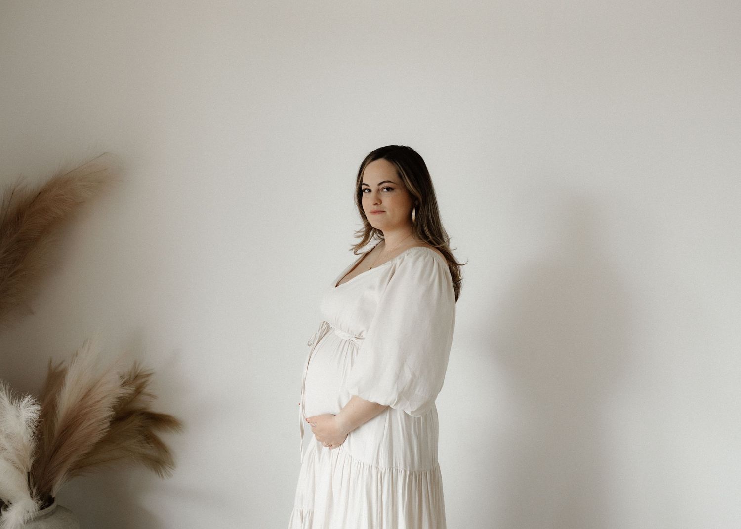 A sequence of images showing a pregnant person in white dress against a neutral wall with pampas grass decoration.