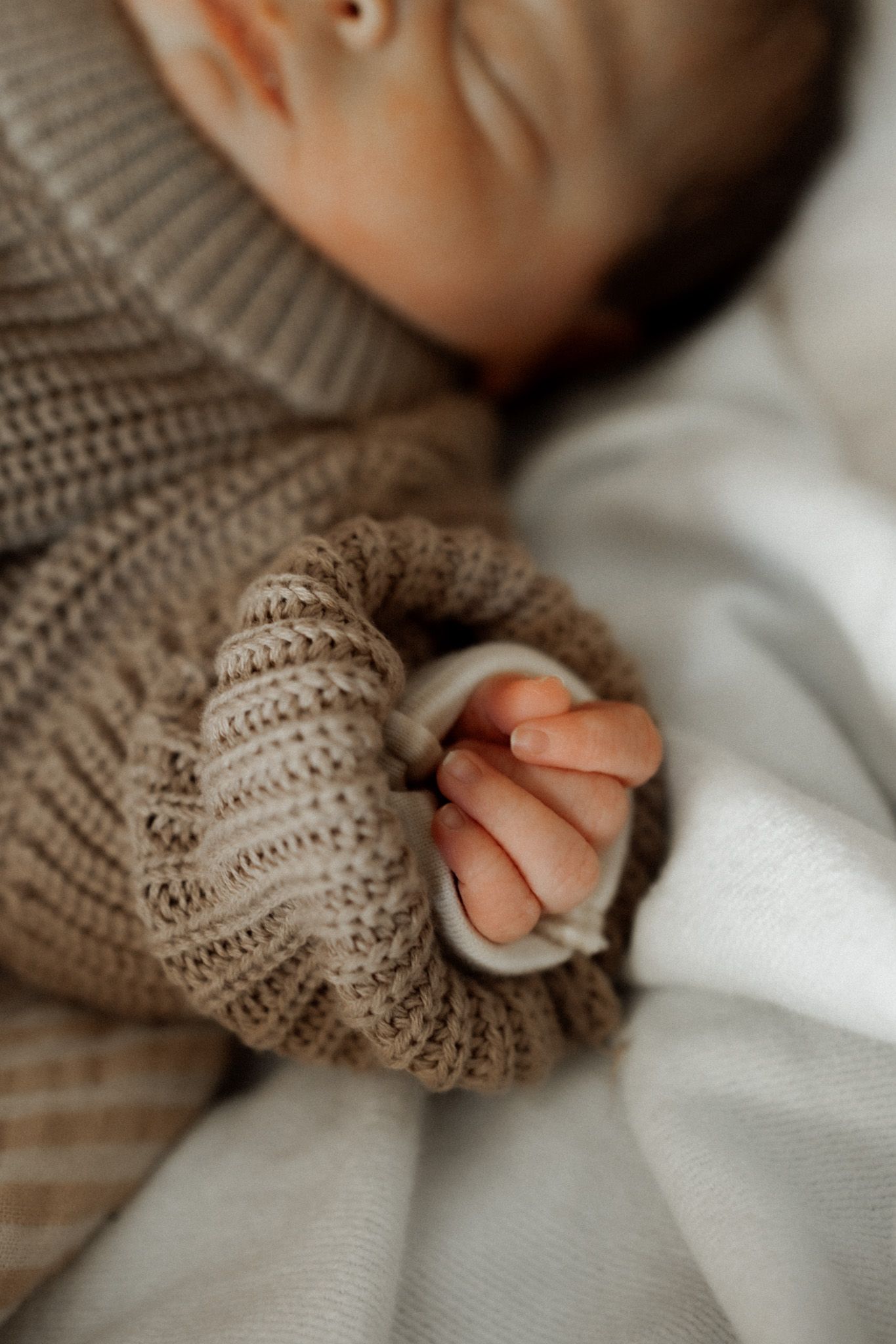 Close-up detail of baby's tiny hand in a knitted brown sweater against white fabric.