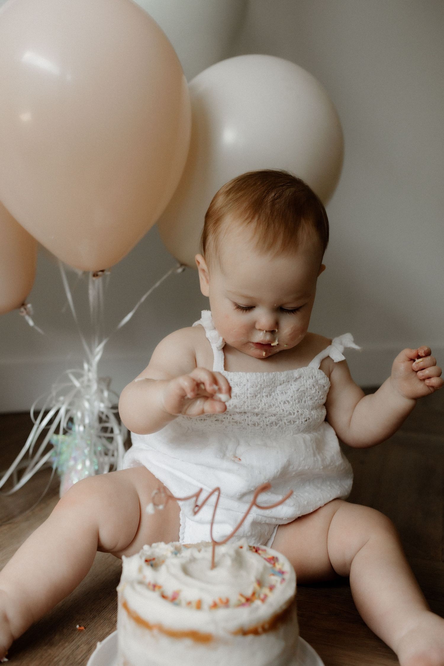 Baby in white dress sitting with first birthday cake and pink and white balloons in background.