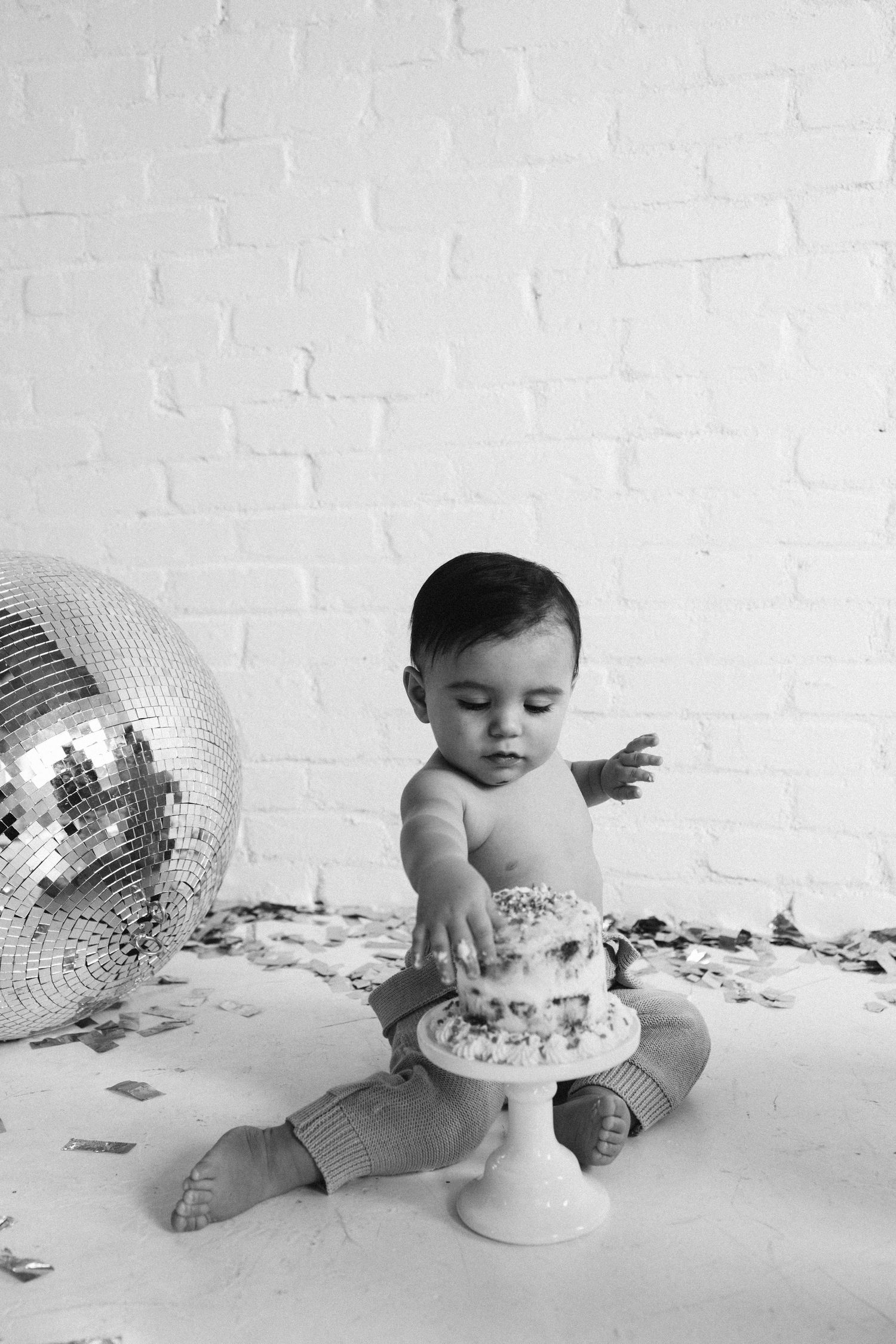 Black and white photo of baby reaching for cake with large disco ball in background.