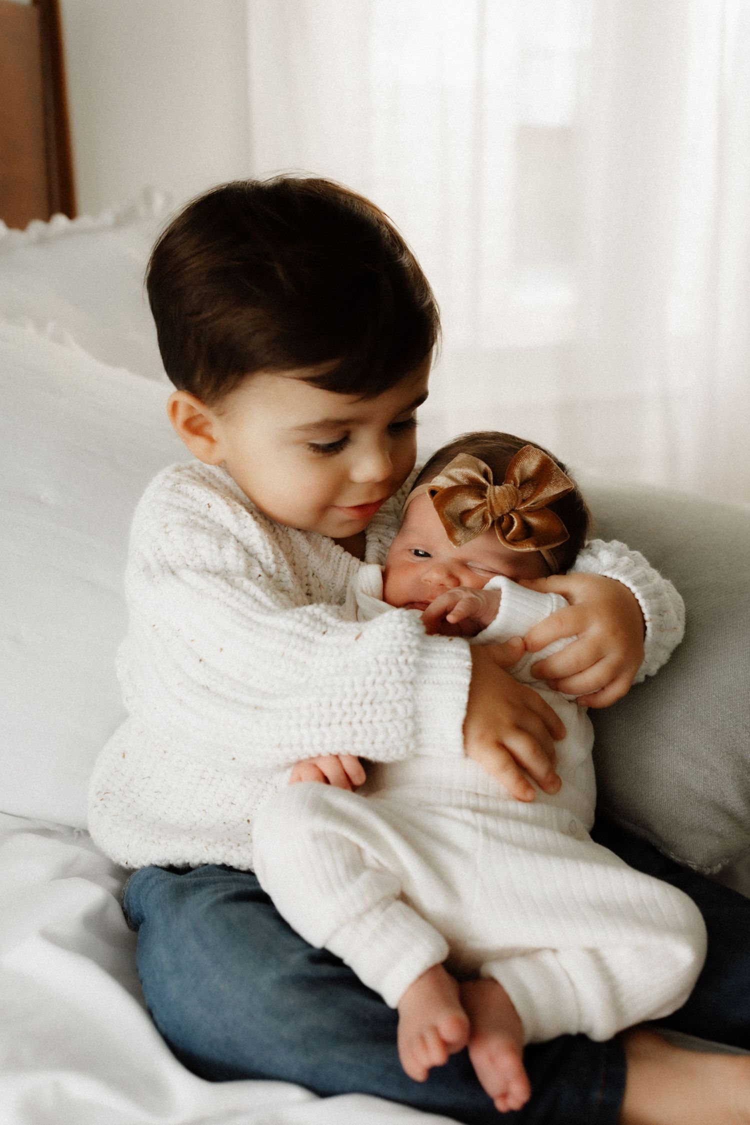 Young child in white sweater gently holding and looking at baby sibling dressed in white.