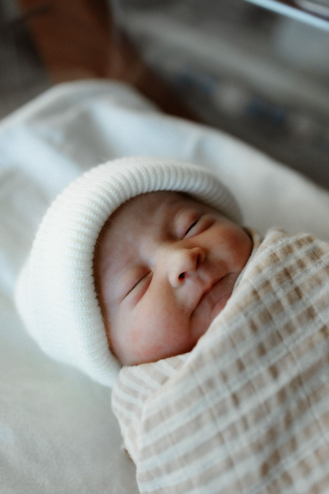 Sleeping newborn wearing a white knit hat and wrapped in checkered blanket.
