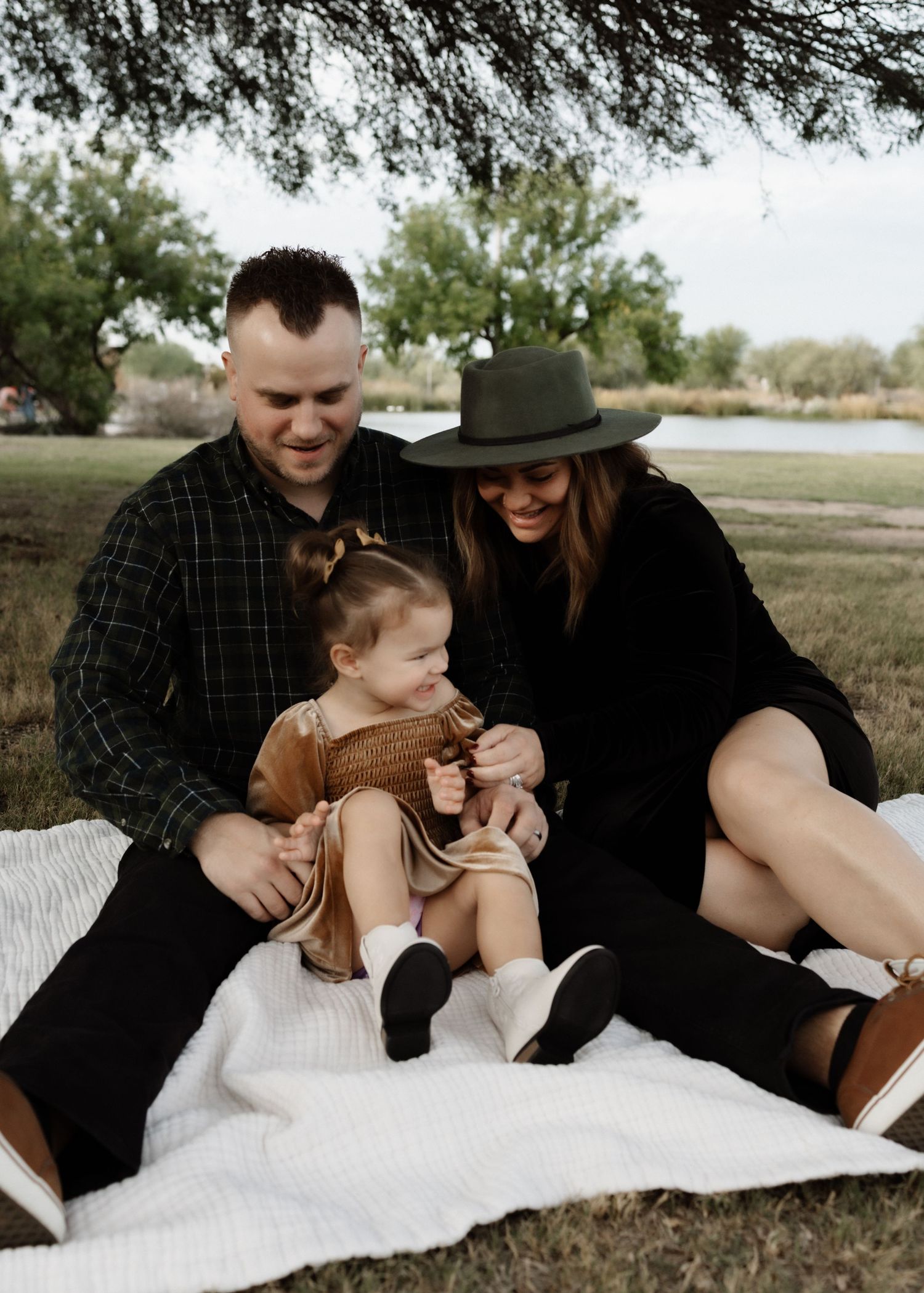 A family enjoys a picnic on a white blanket under a tree near a lake.
