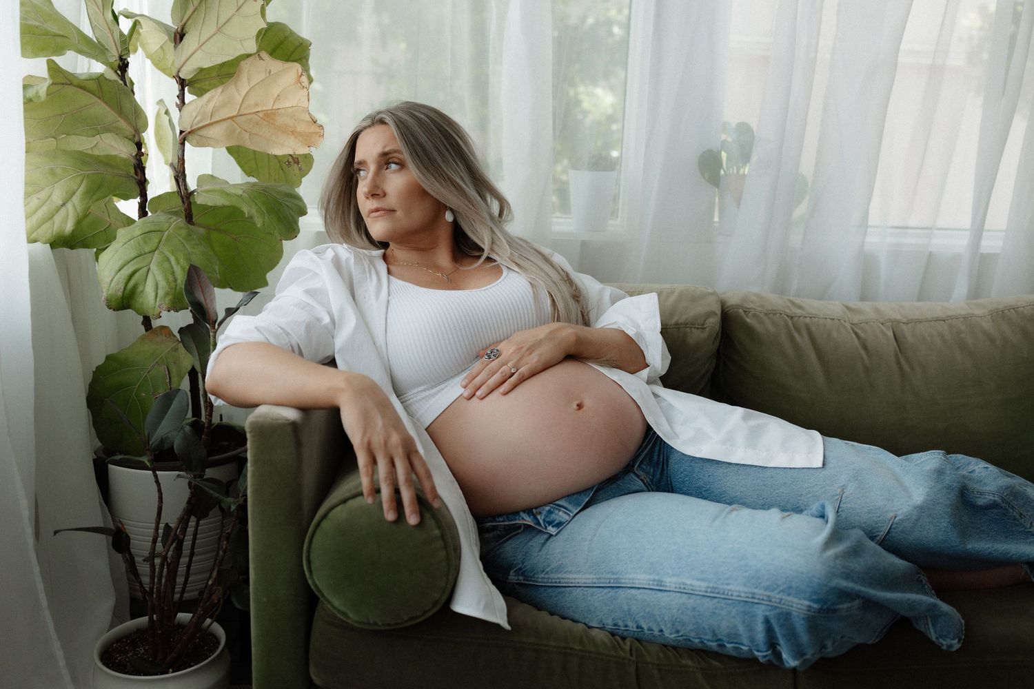 Expectant mother rests comfortably on blue couch near window with sheer curtains and indoor plant.