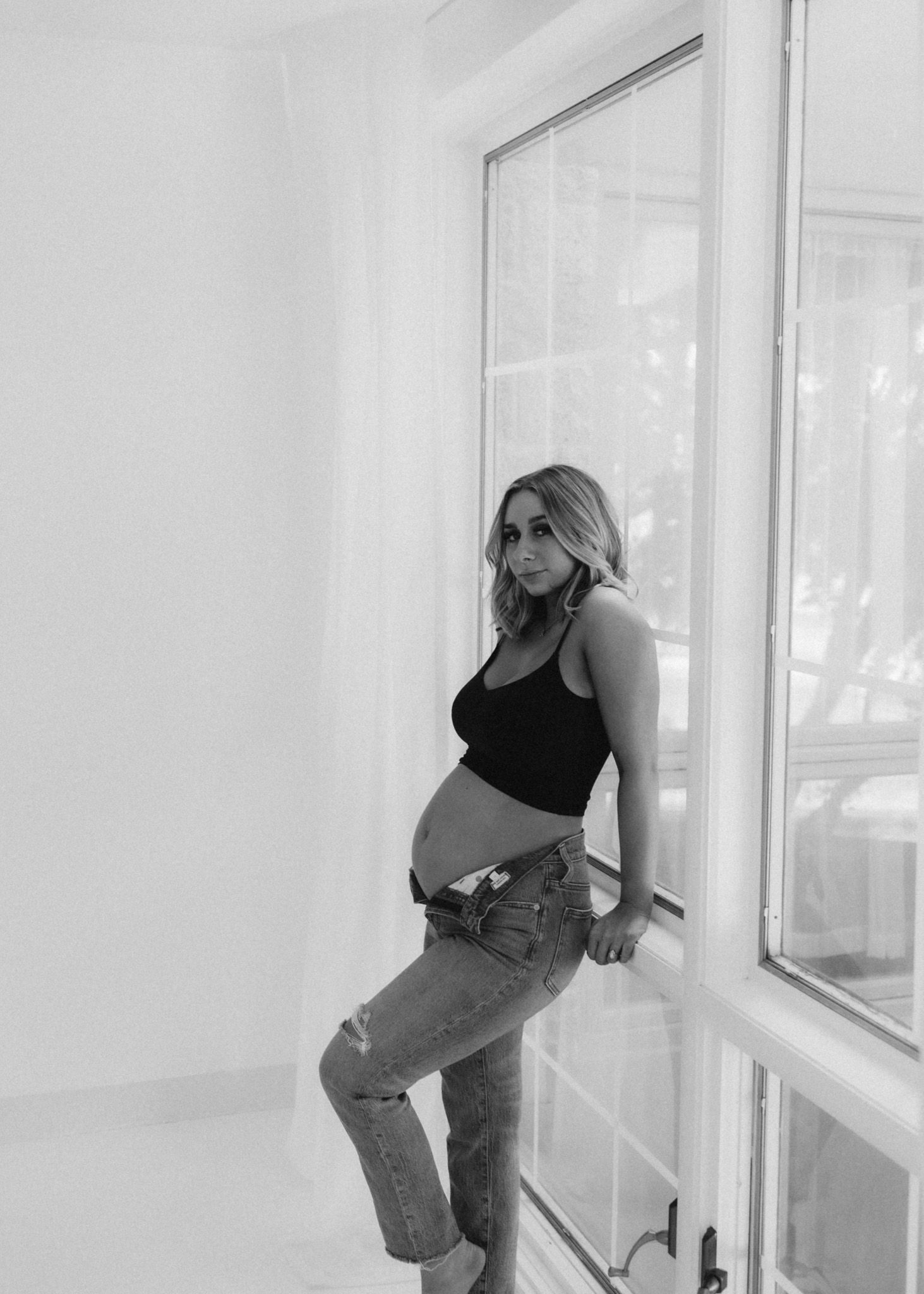 Artistic black and white maternity portrait by window in casual denim.