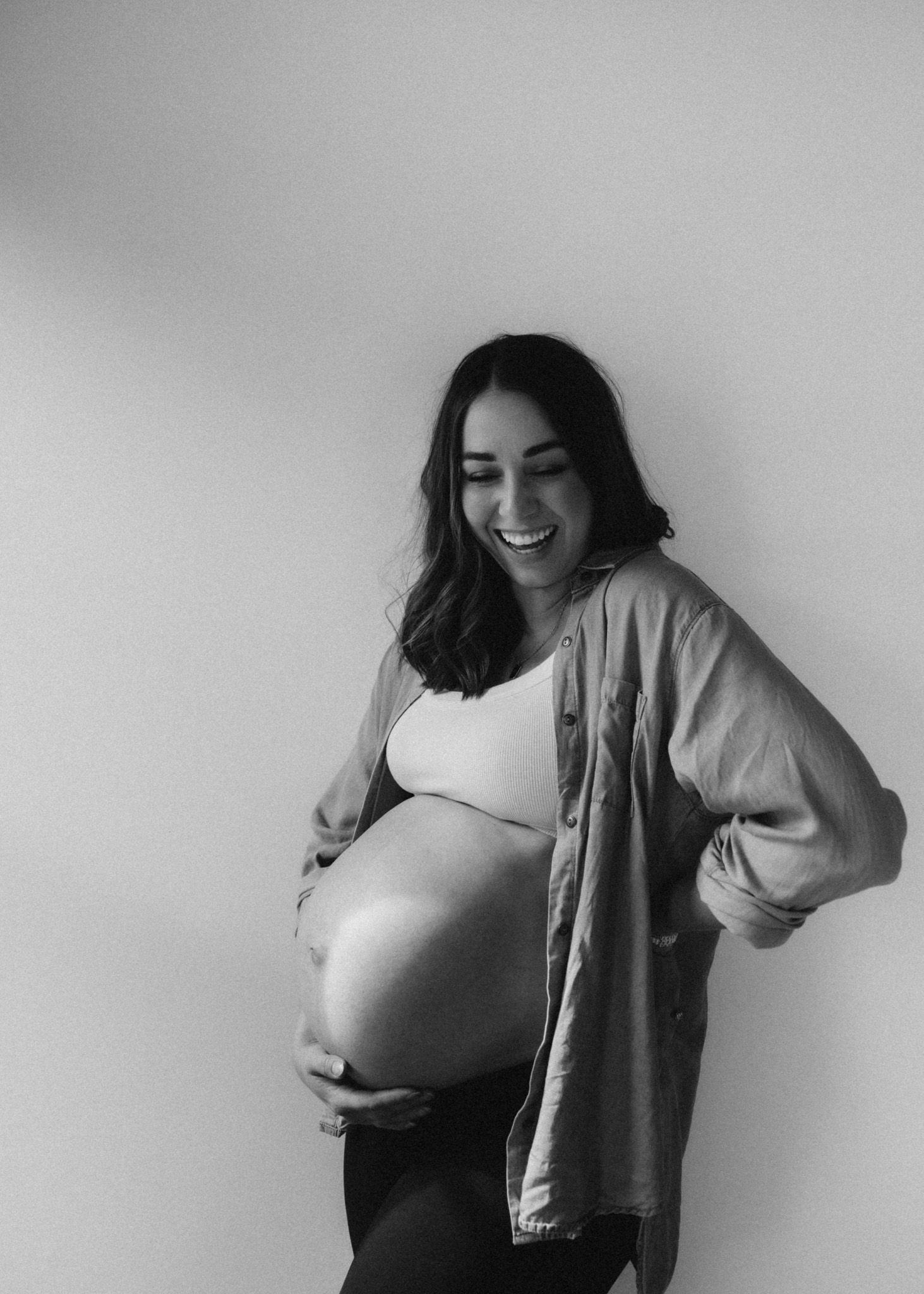 Black and white portrait of pregnant woman in oversized shirt smiling against wall.