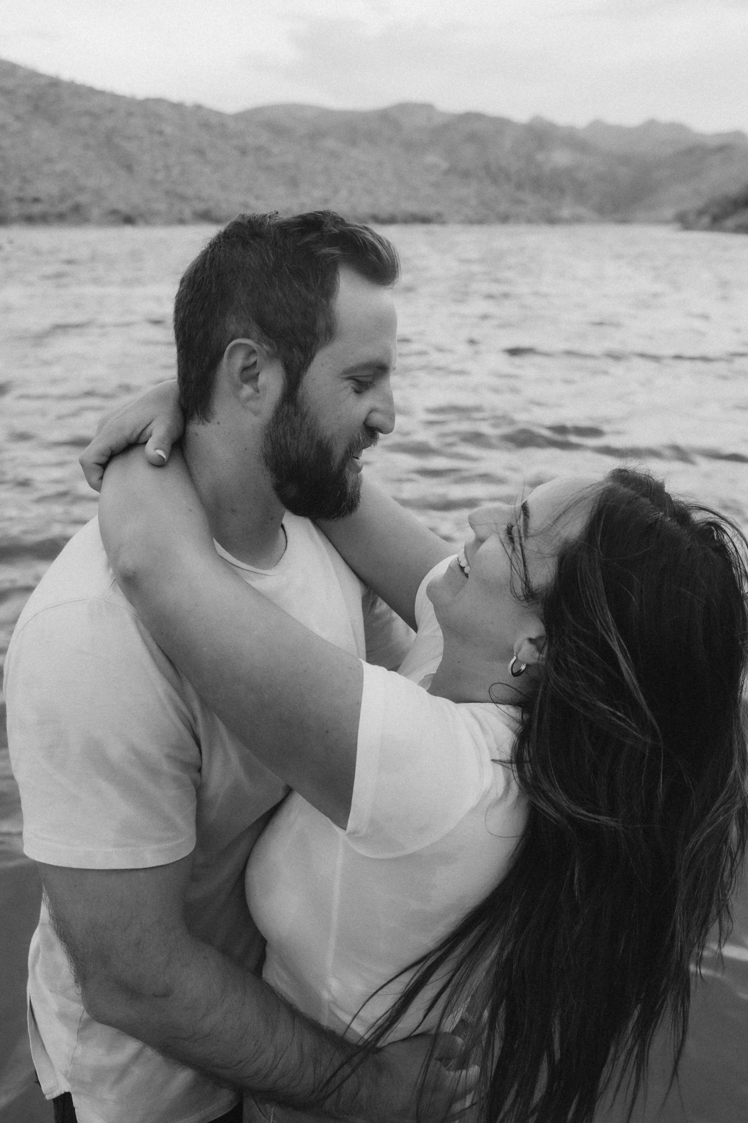 A couple embraces passionately by the water with mountains in the background, shown in black and white photos.