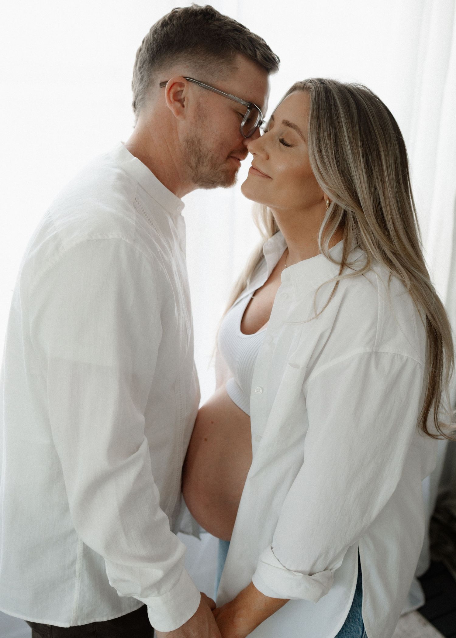 Intimate couple in matching white shirts share a tender embrace by a bright window.