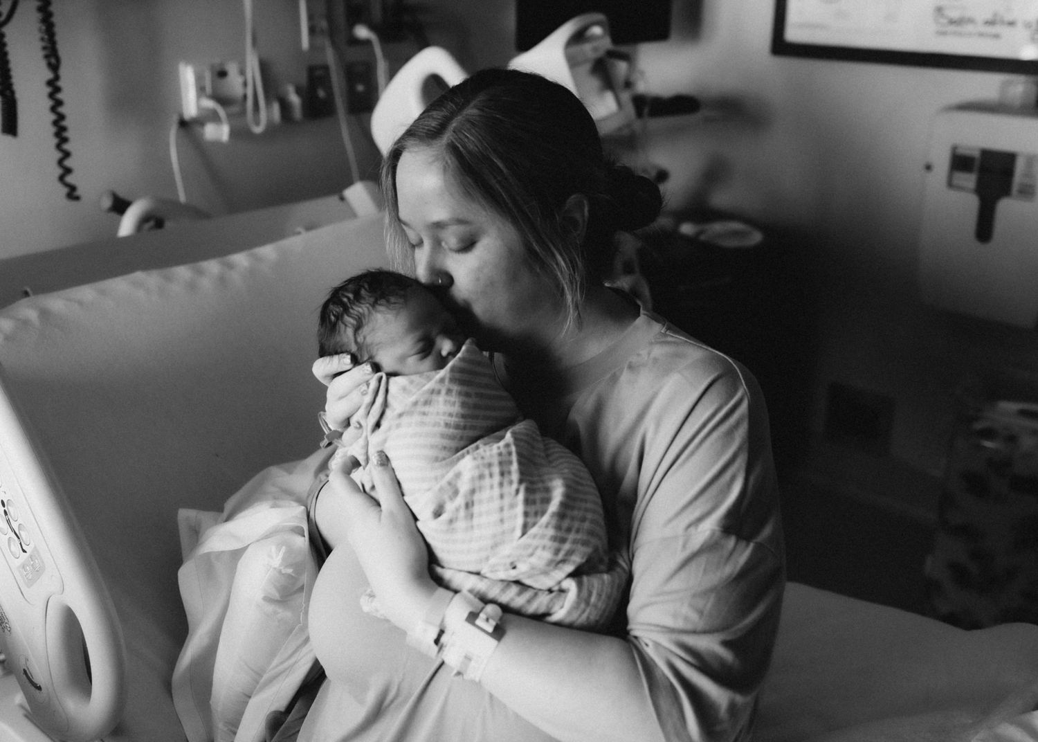 Black and white image showing a tender moment between mother and newborn in a hospital room.