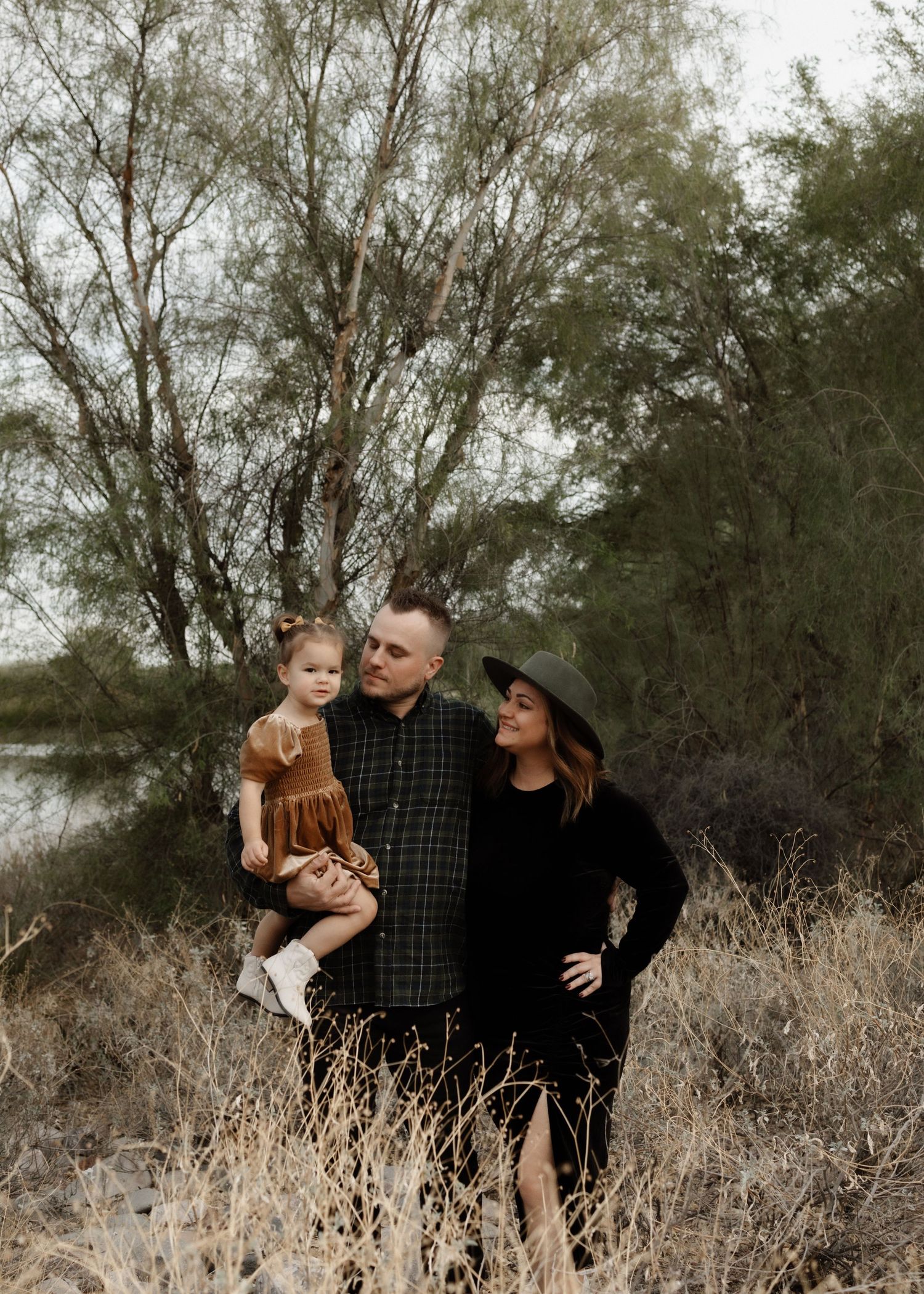 A family poses together outdoors with tall trees and a lake in the background.