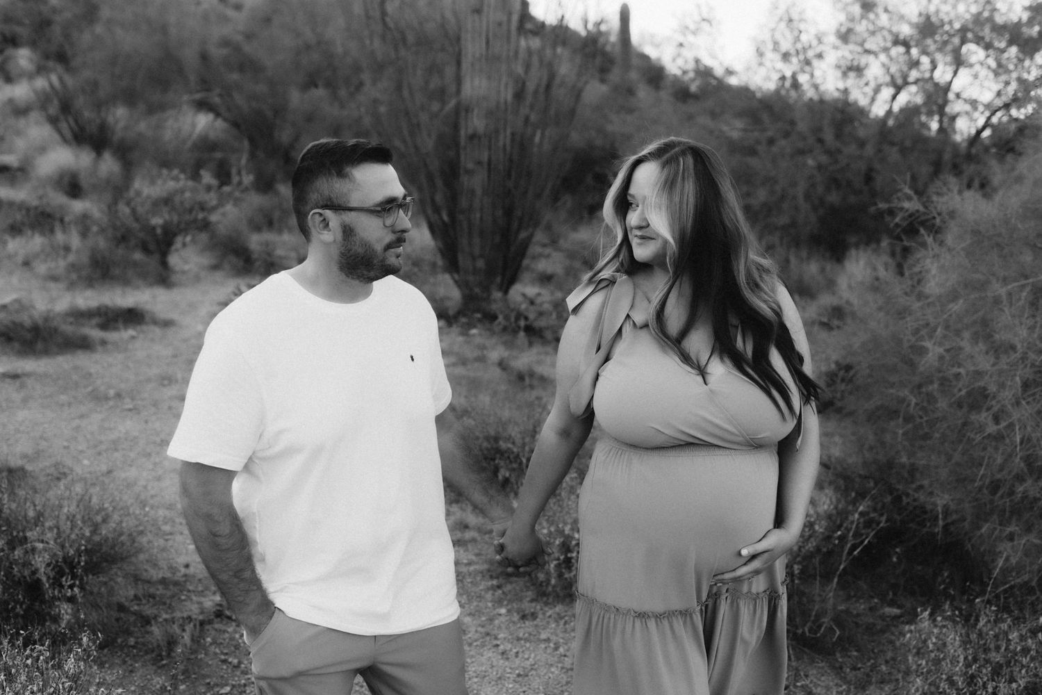 A black and white photo of an expectant couple smiling together in a desert landscape with saguaro cacti.